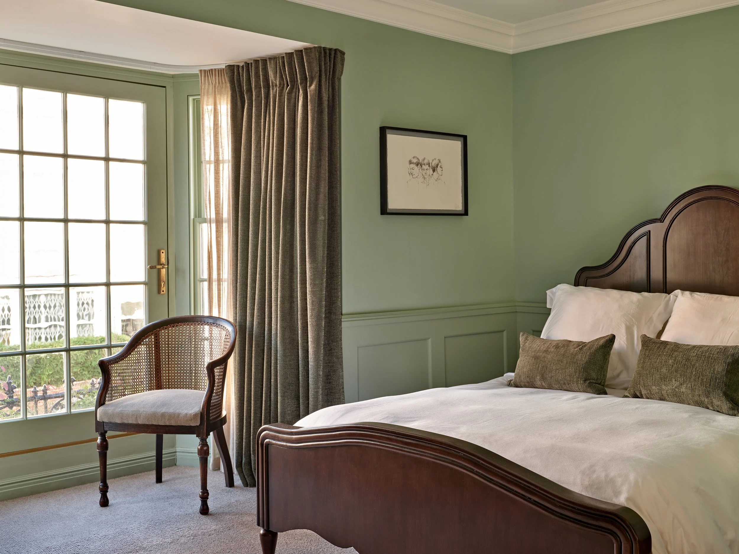 A bedroom with a large window and curtains, a framed picture on the wall, a bed with white linens and pillows, a wooden headboard, and a patterned chair near the window.