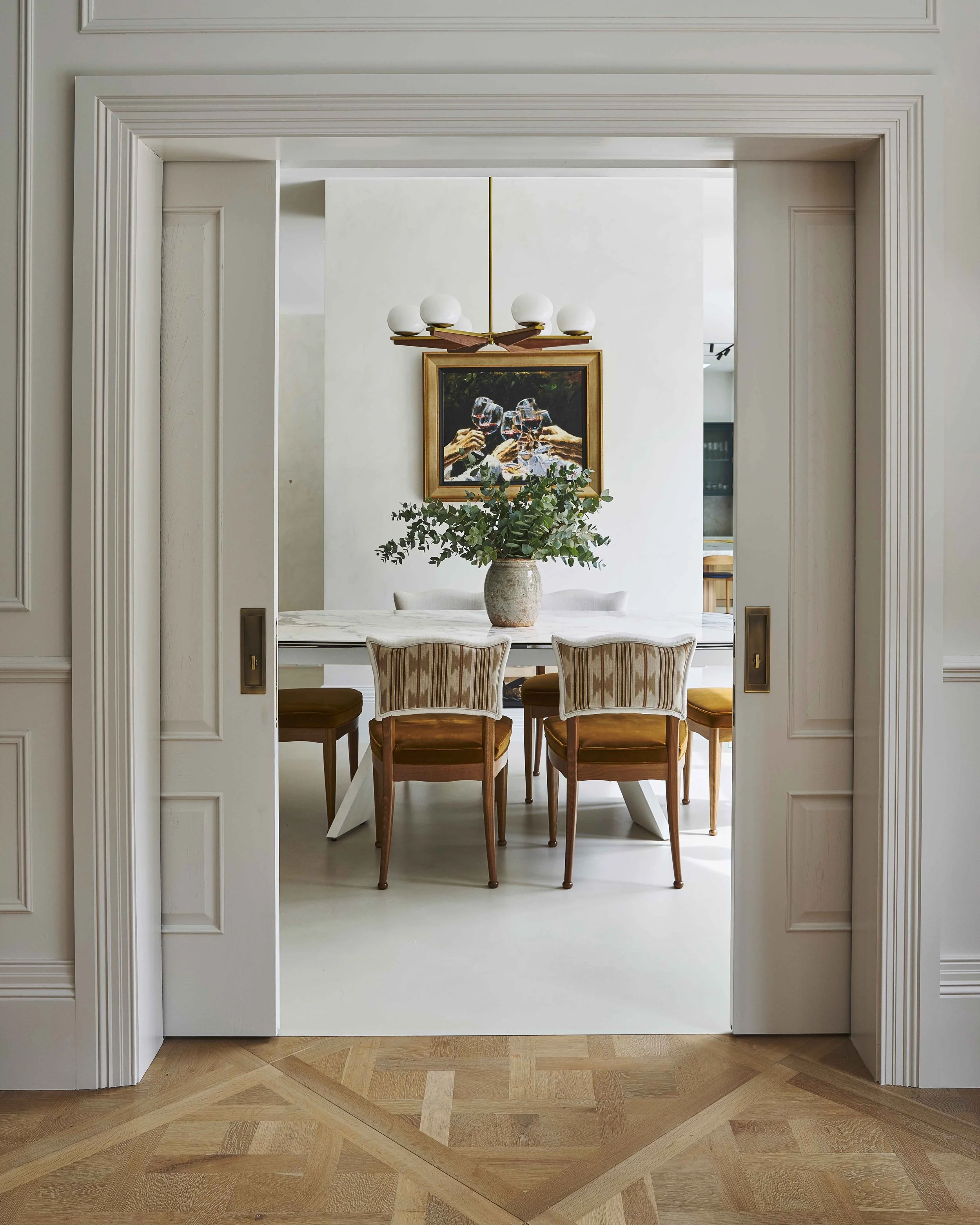 A view through an open doorway into a dining room, which features a table with a floral centerpiece, surrounded by chairs, a painting on the wall, and an elegant chandelier hanging from the ceiling.
