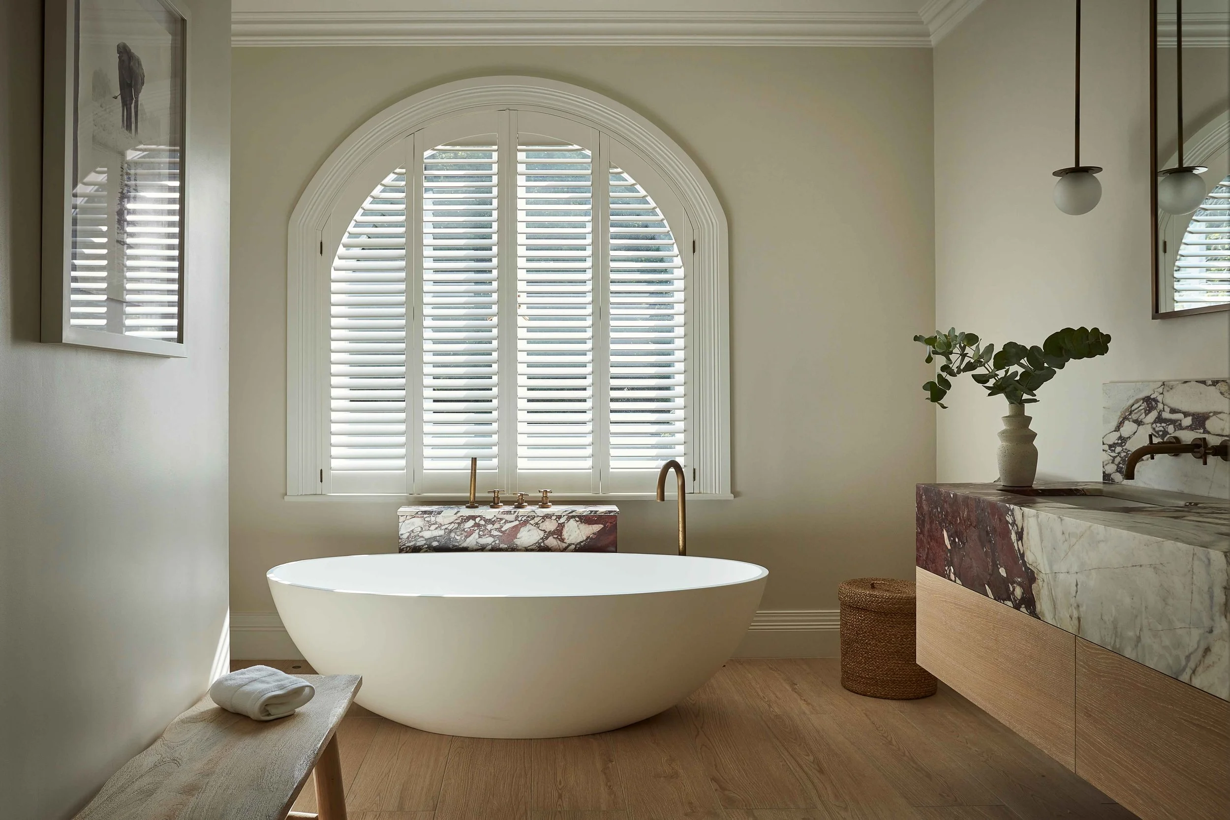 A modern bathroom with a large arched window with white shutters, a freestanding bathtub, a marble countertop, a potted plant, and minimal decor.