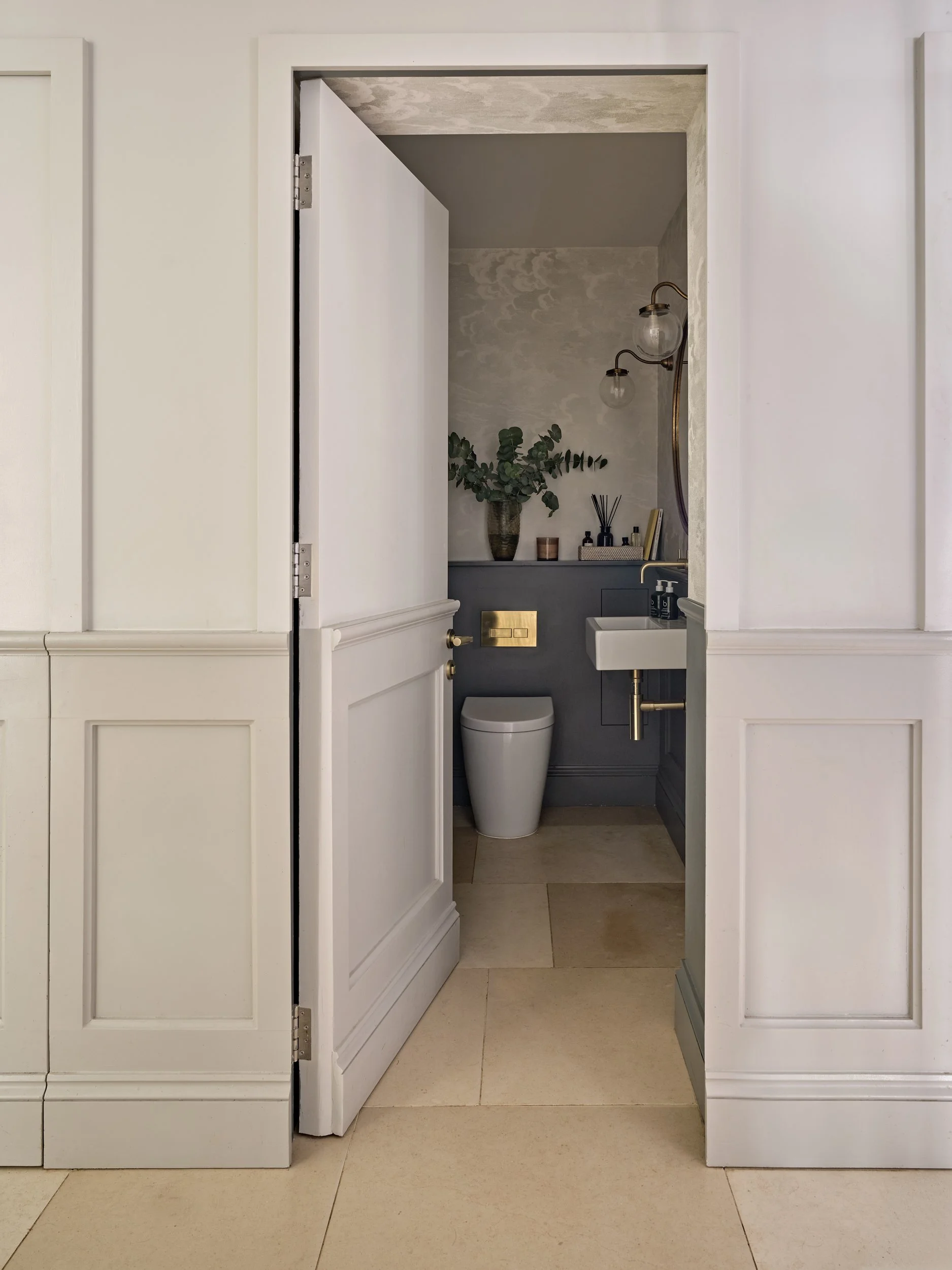 A bathroom with white cabinets, a small white sink, a toilet, a black cabinet with a gold flush button, a potted plant, diffuser, and a wall lamp with exposed light bulbs.