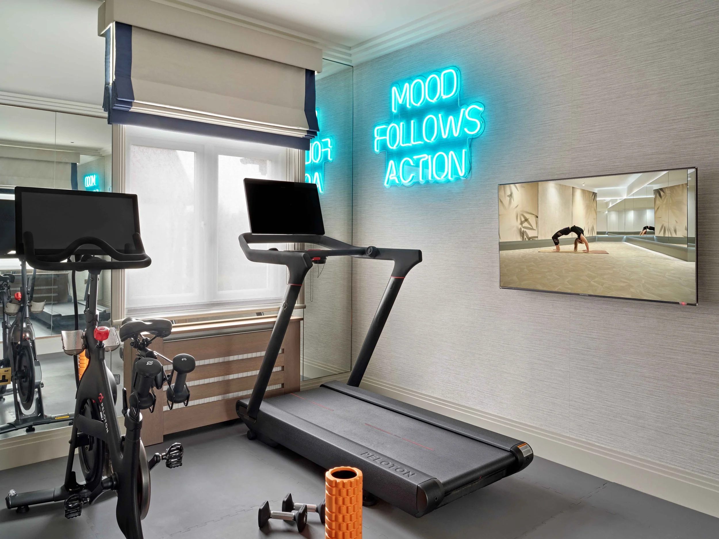 Home gym with treadmill, exercise bike, and a neon sign that says 'Mood Follows Action' on the wall. A mirror and window are also visible.