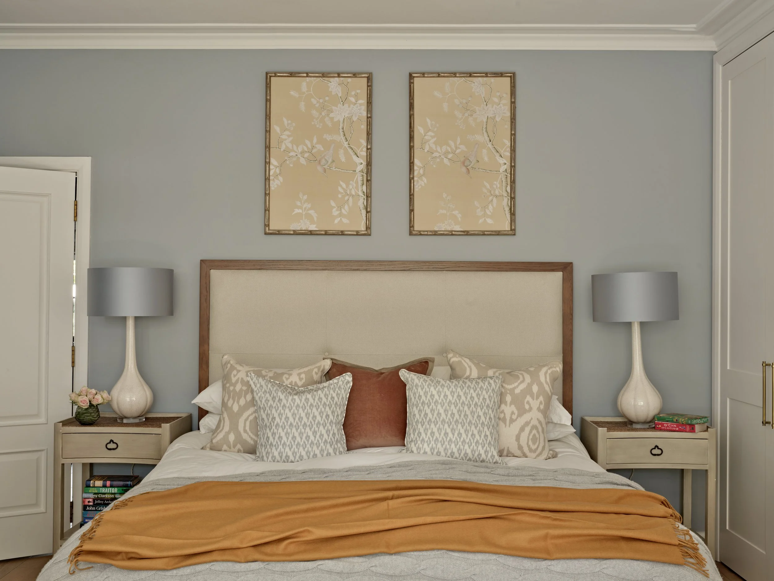A bedroom scene with a bed, multiple pillows, and two matching bedside tables with lamps. Framed artwork of bird and branch motifs hangs above the bed on a gray wall.