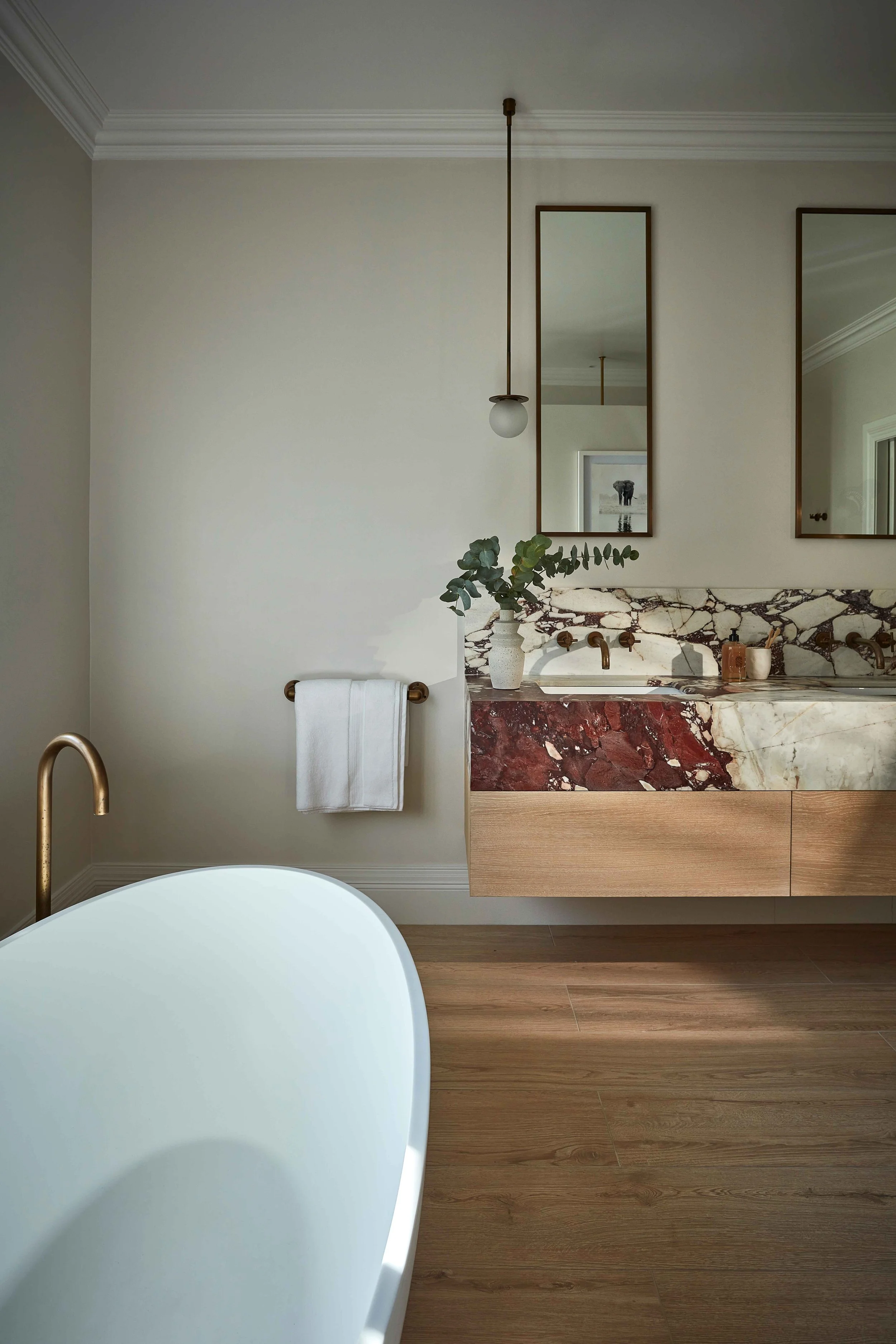 Bathroom with a white freestanding bathtub, modern wooden flooring, a wall-mounted marble vanity with gold fixtures, a potted plant, and a large mirror.