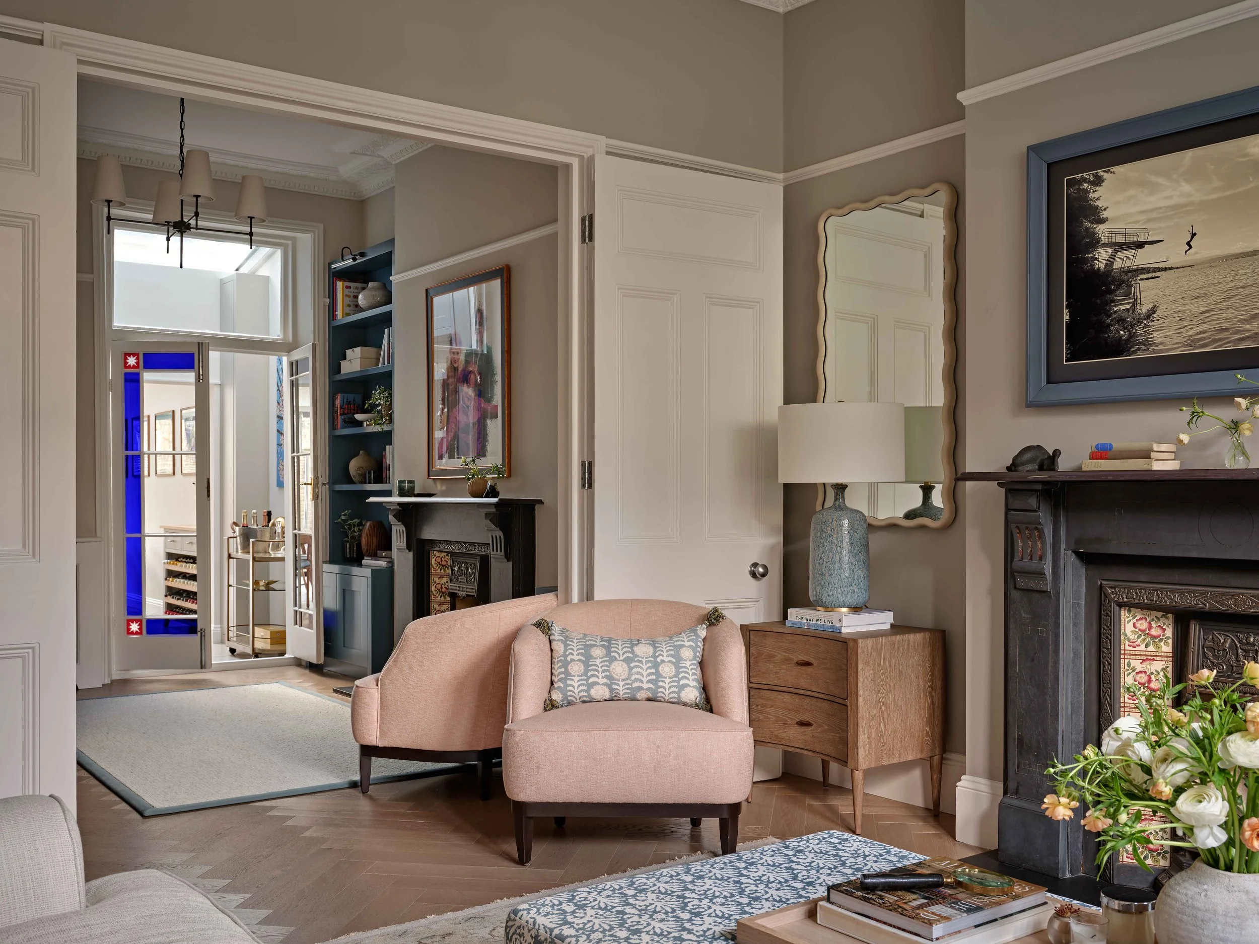 Interior view of a living room with a pink armchair, a side table with flowers and books, a fireplace, and framed pictures on the wall.