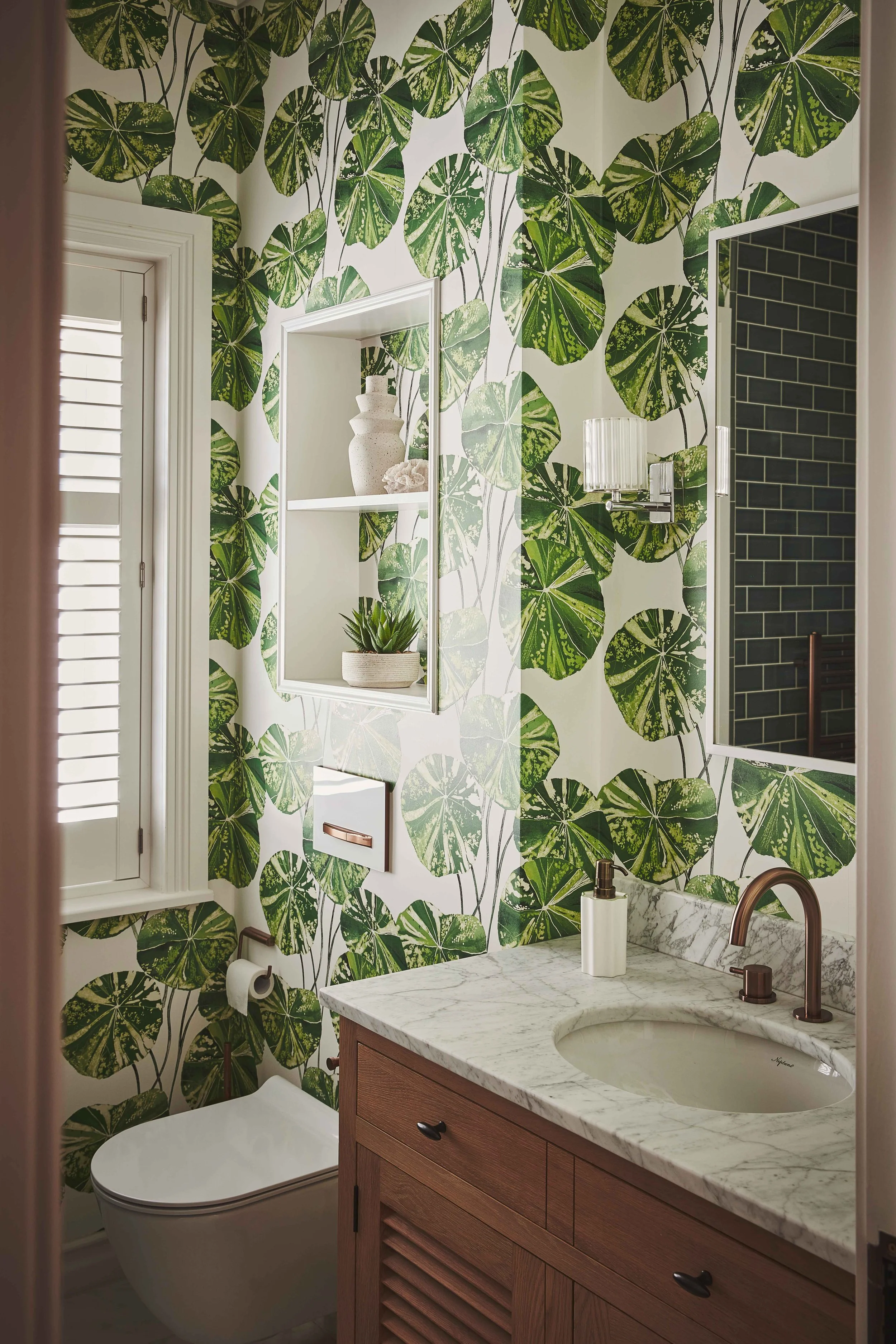 Bathroom with green leafy wallpaper, white window shutters, decorative shelf with vases and plant, marble countertop with sink, and a mirror.