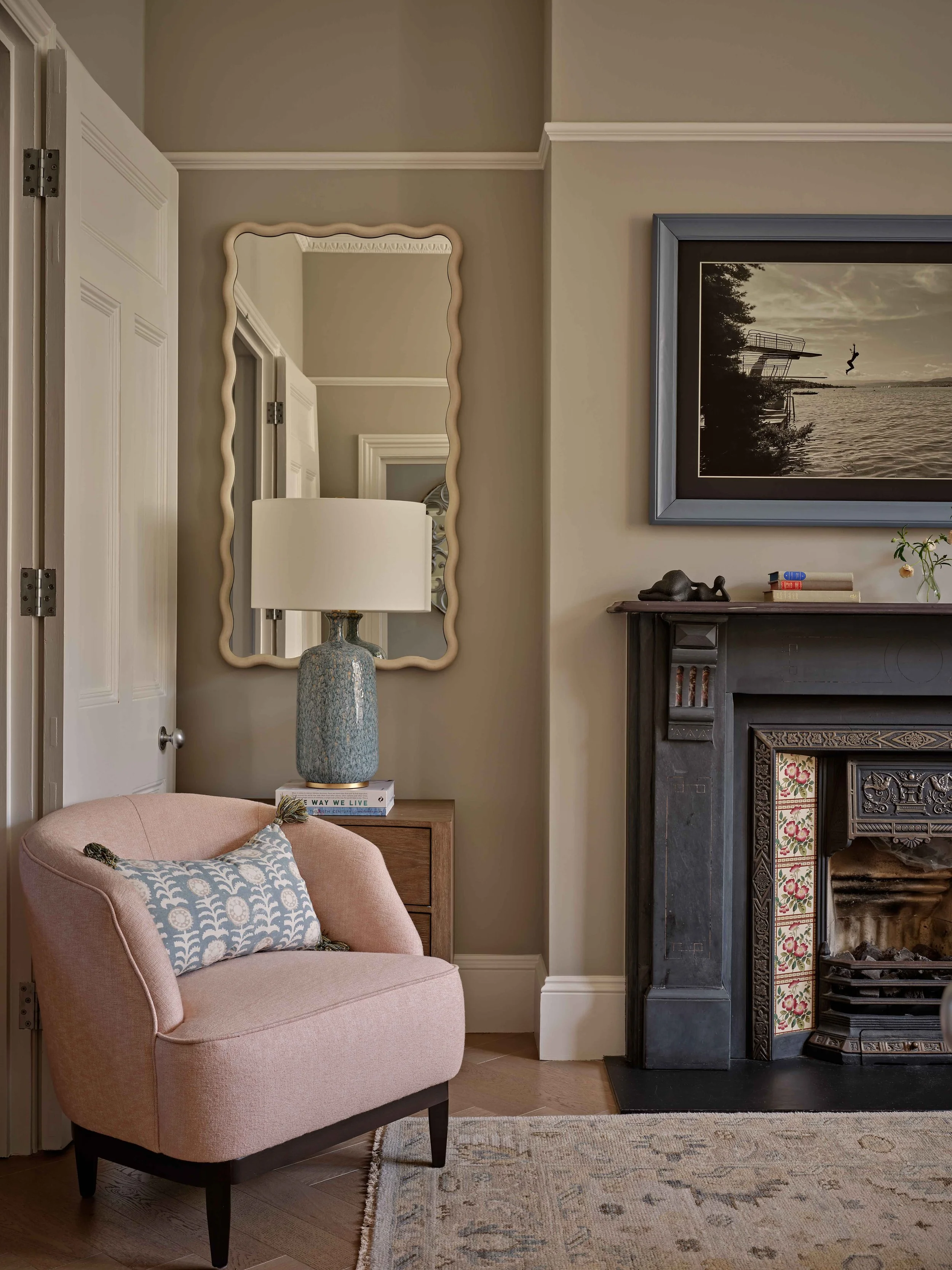 A cozy living room corner displaying a pink armchair with a decorative pillow, a wooden side table with a ceramic table lamp, a large wall mirror with a wavy frame, a framed black and white photo of a diver jumping into water, and a dark fireplace wi