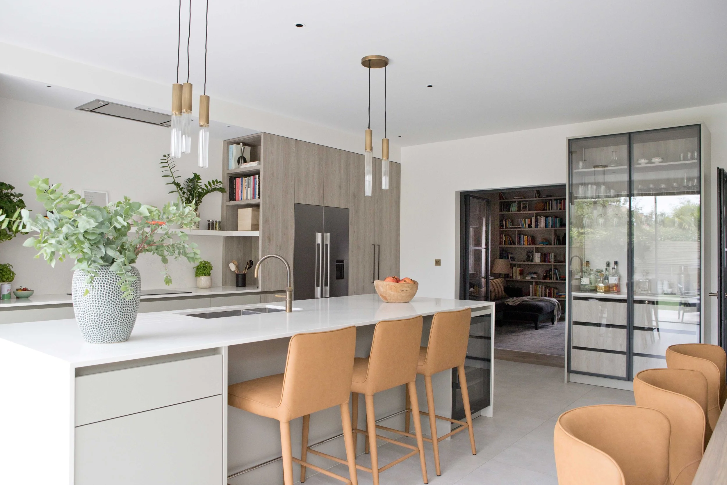 Modern kitchen with white island, tan chairs, green plants in a white vase, gray cabinets, and a glass door leading to a room with bookshelves.