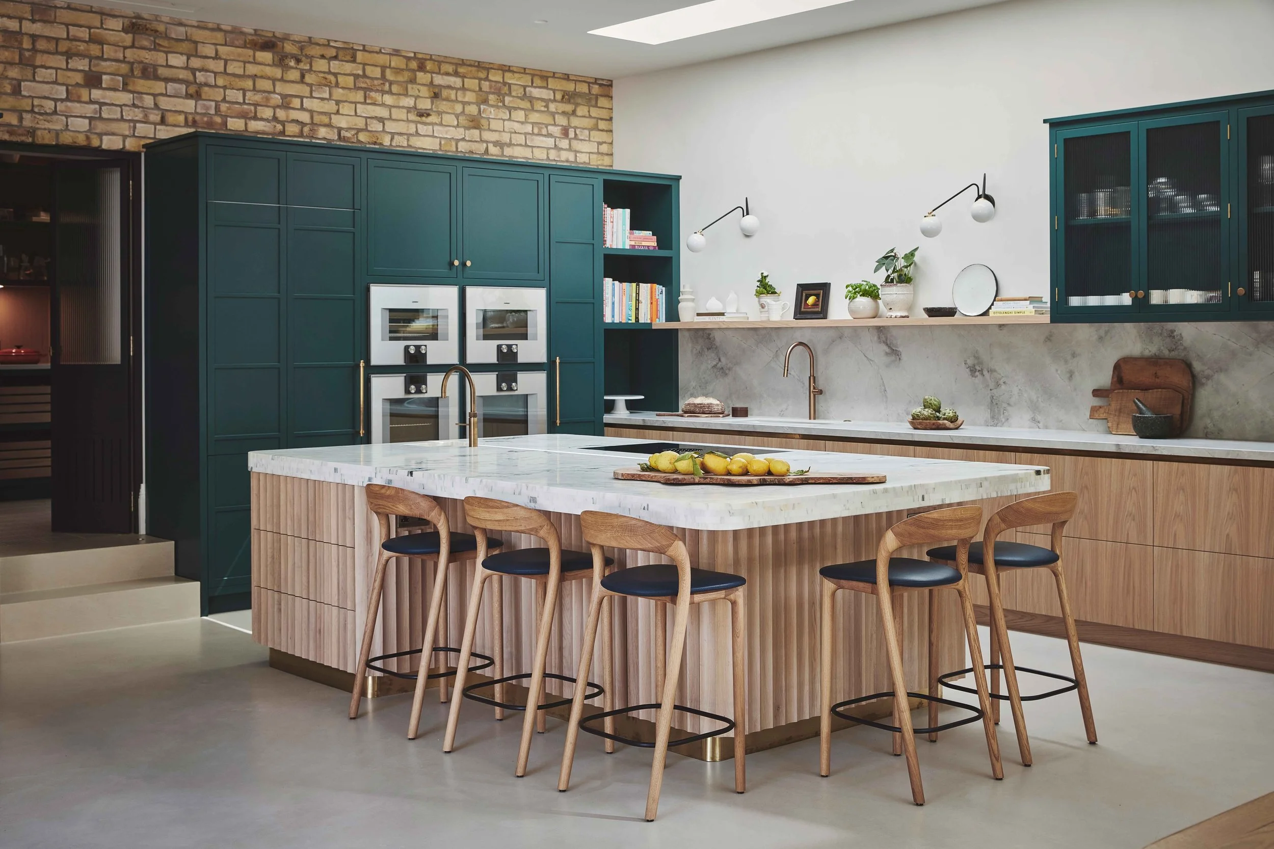 Modern kitchen with a large white marble island, teal cabinetry, open bookshelves, and exposed brick wall. Kitchen decor includes potted plants, ceramic vases, and minimalist lighting.