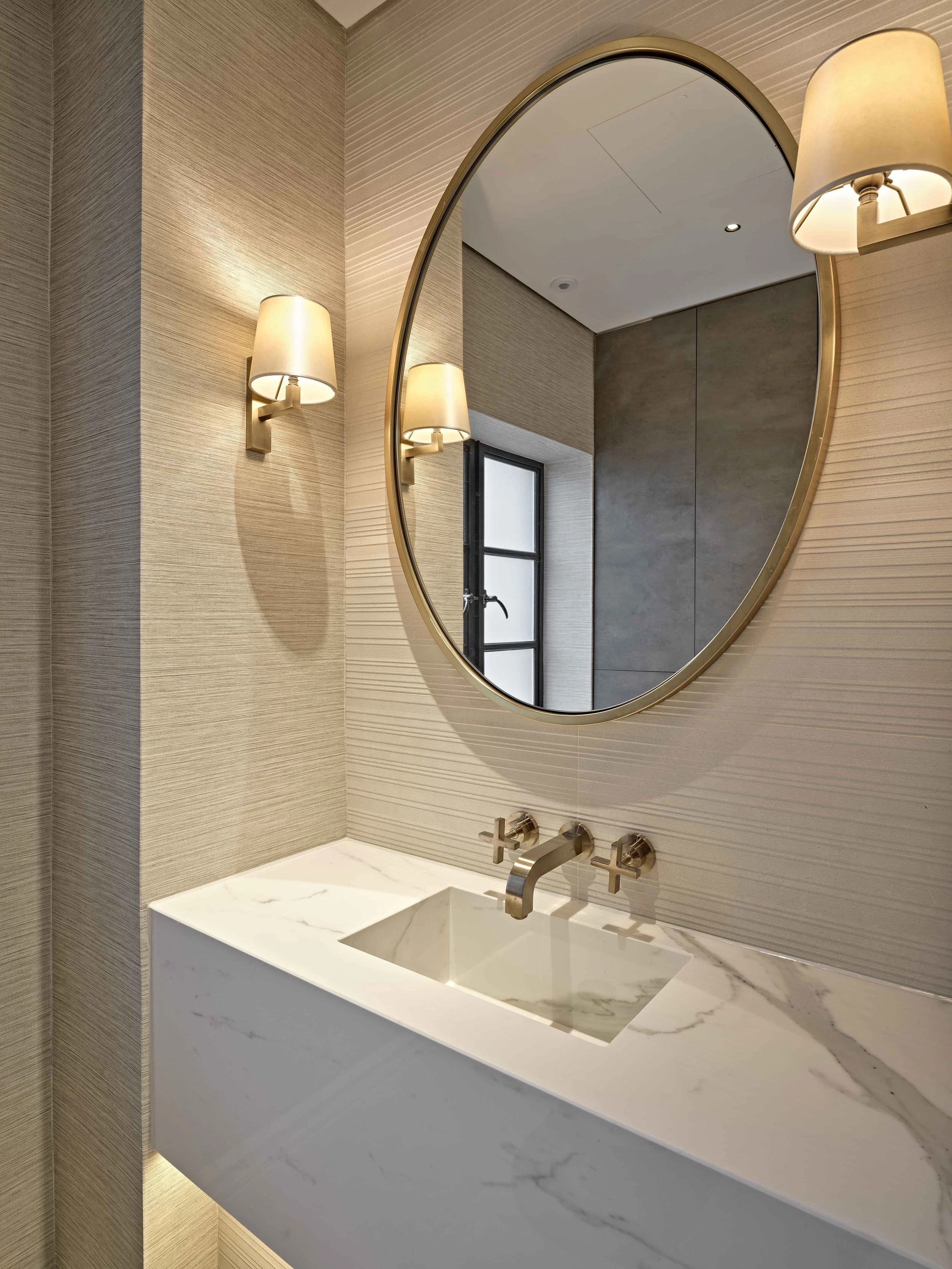 A modern bathroom sink with a marble countertop, a wall-mounted faucet, a large round mirror, and two wall-mounted lamps with beige shades, beige textured wall panels, and a small window
