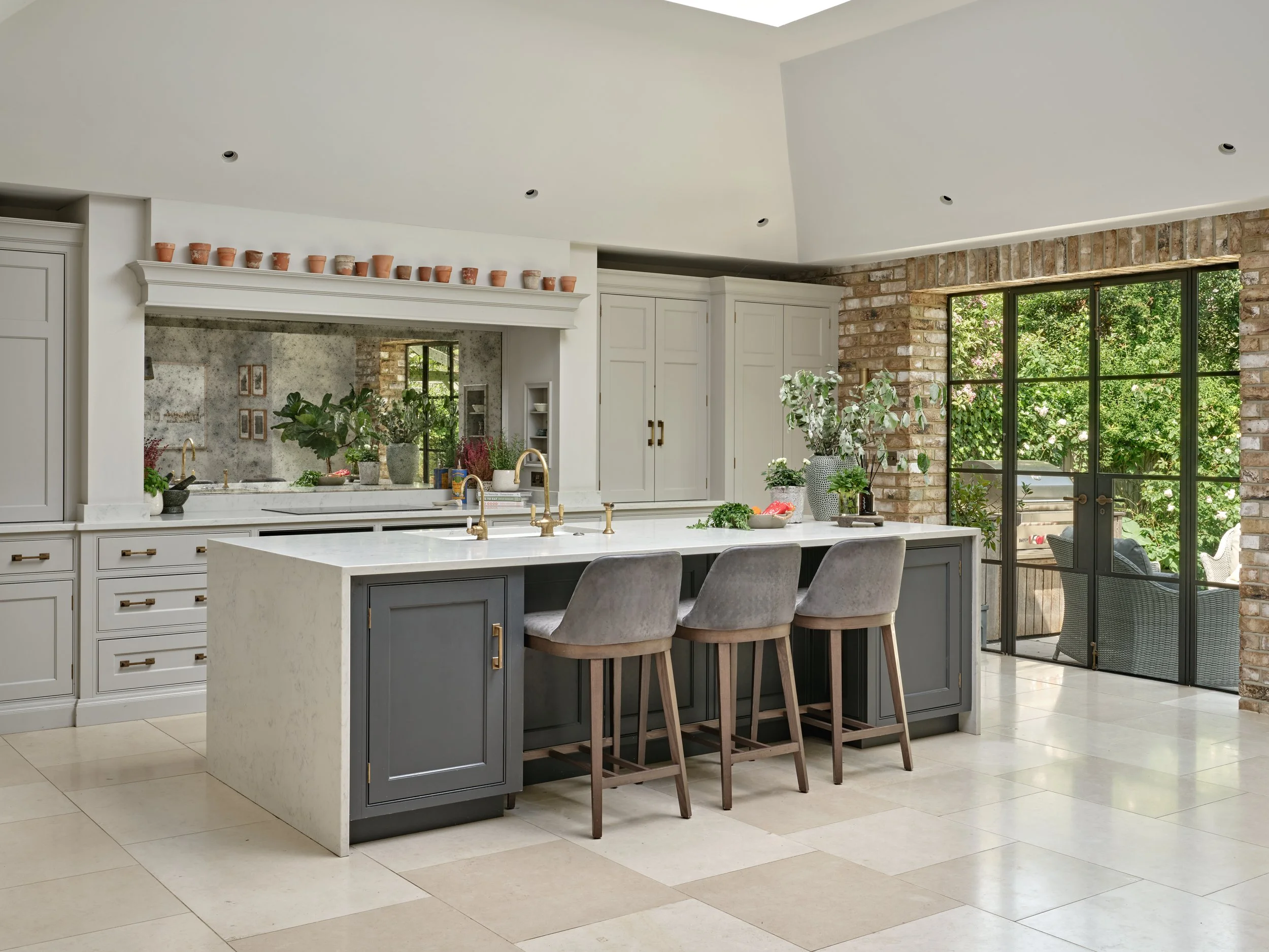 Modern kitchen with white cabinetry, a large island with a white marble countertop, gray chairs, a brick accent wall, large windows with greenery outside, and potted plants.