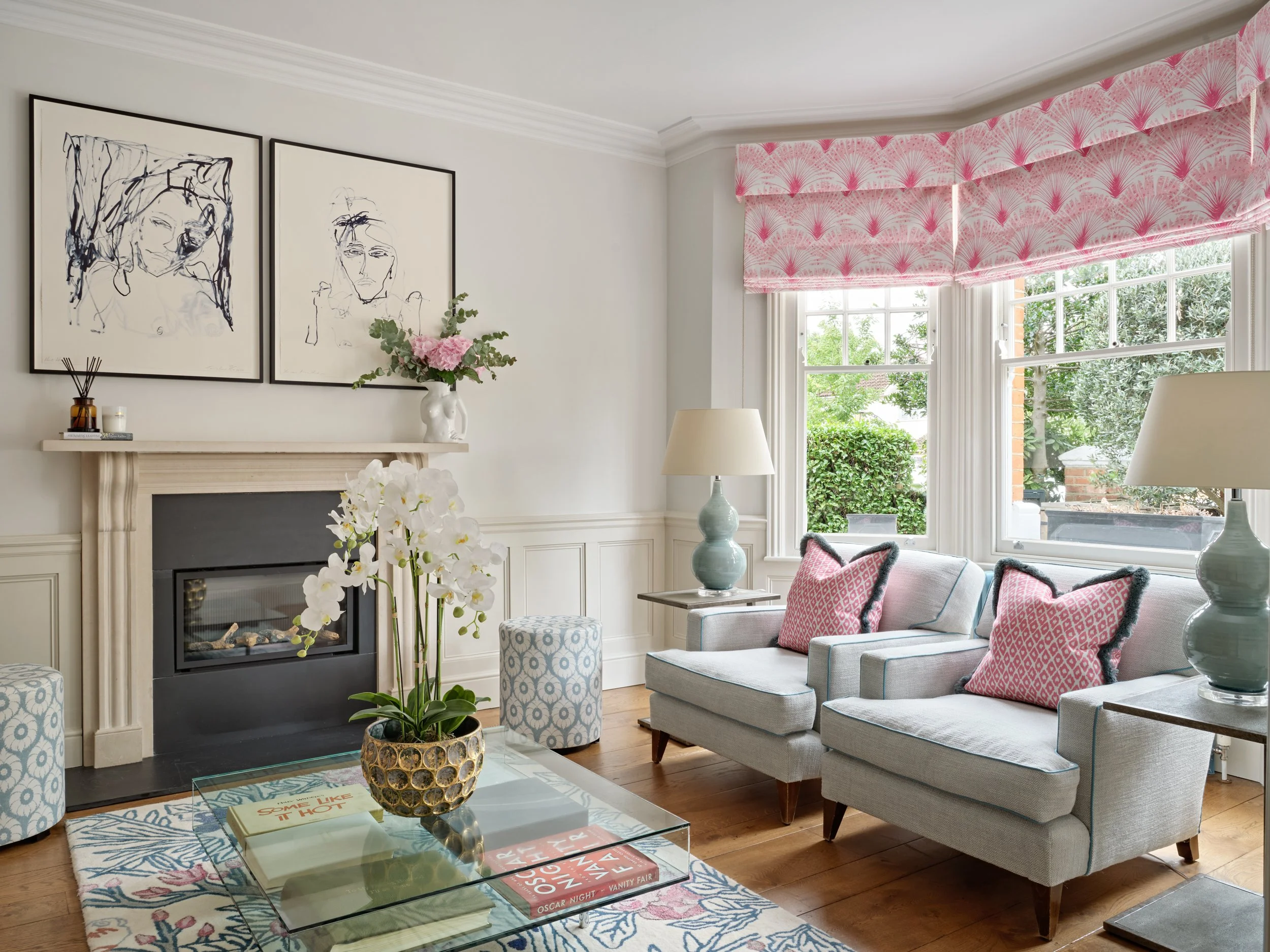A bright living room featuring a fireplace with black trim, framed abstract line art portraits above it, and a mantel decorated with pink flowers in a white vase, candles, and books. Large windows with pink patterned Roman shades allow natural light 