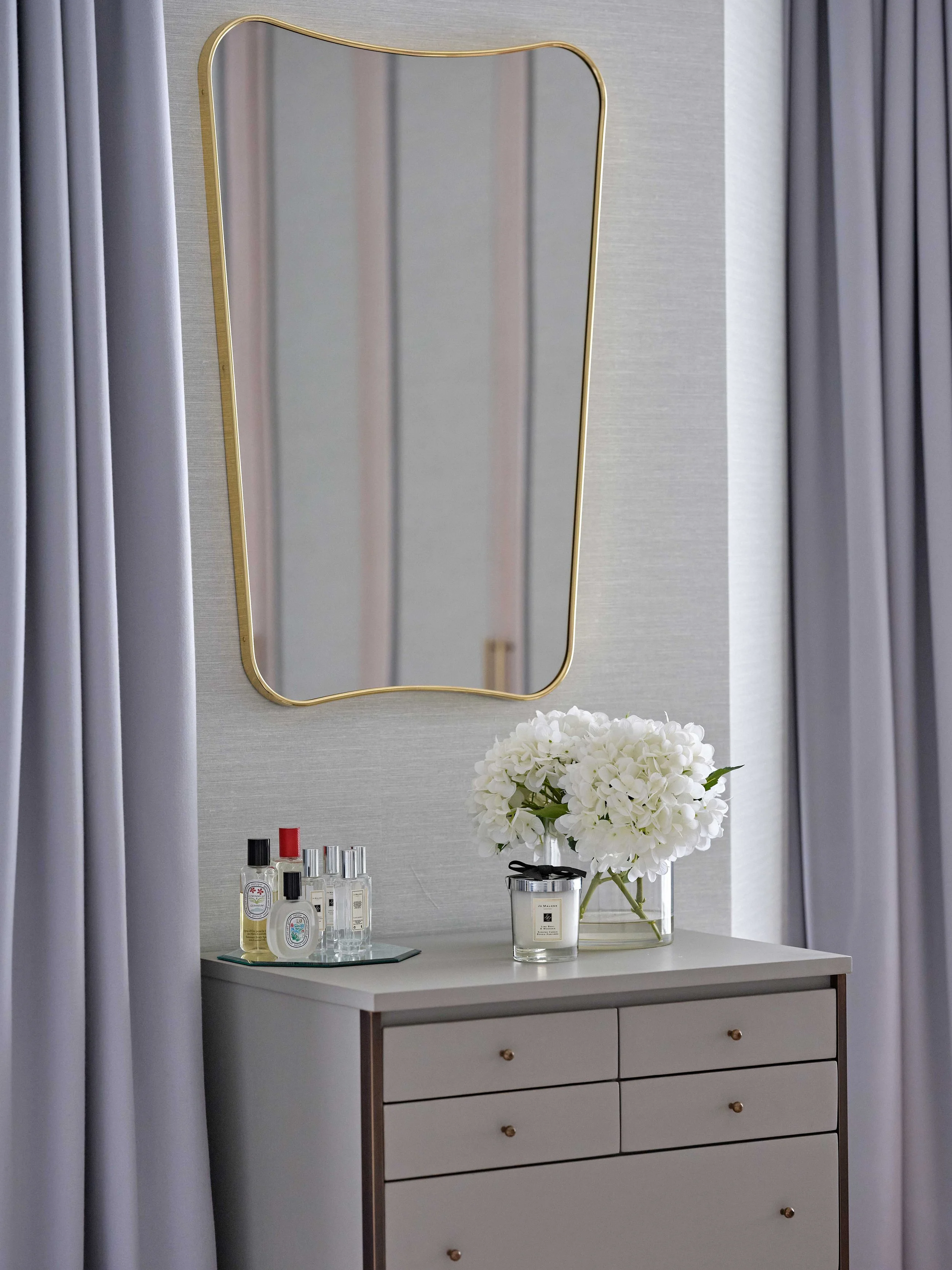 A modern dresser with a white surface and four drawers, topped with a glass vase of white hydrangeas, several perfume bottles, and a Jo Malone candle, with a gold-framed mirror and gray curtains in the background.
