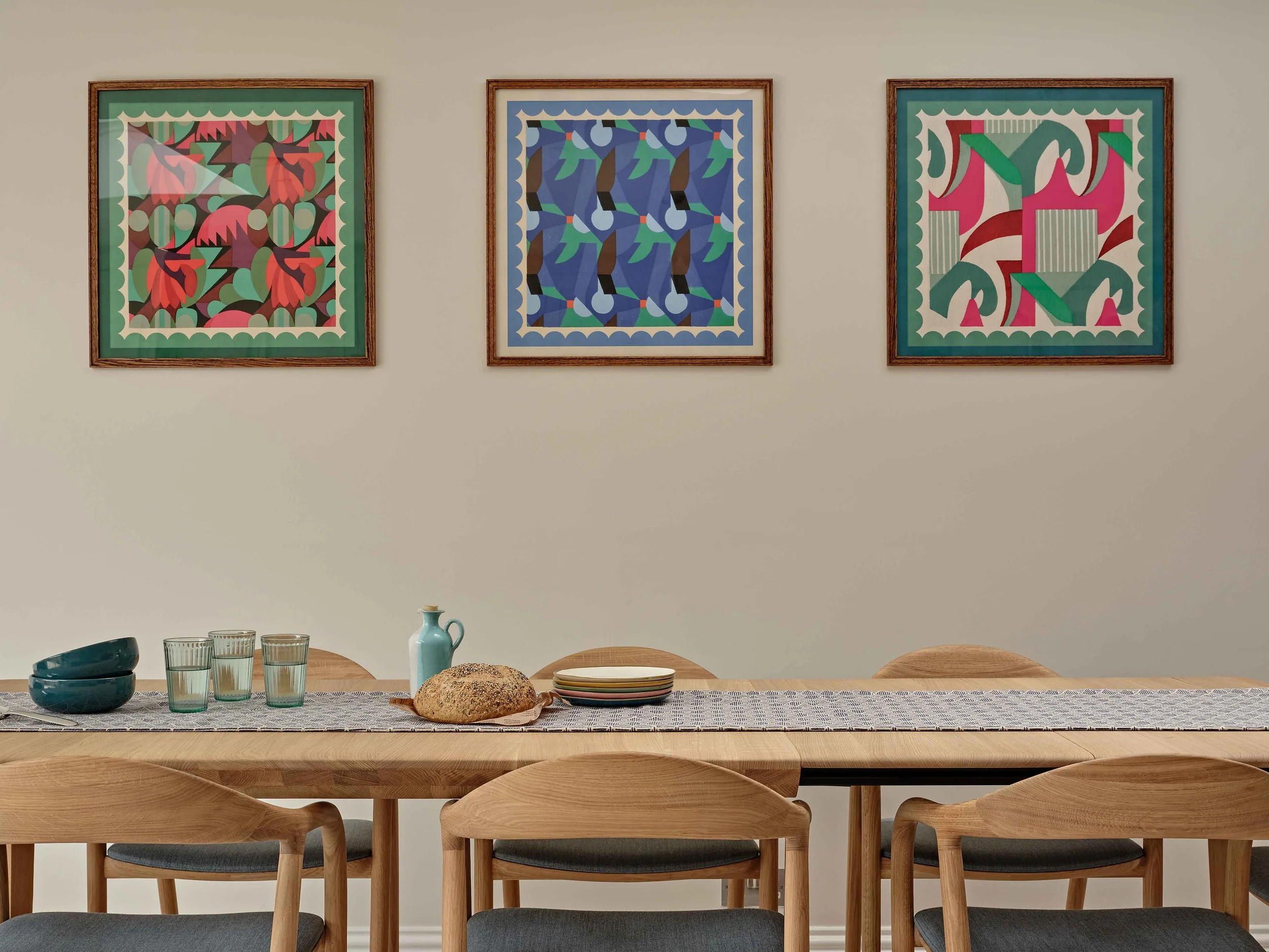 A wooden dining table set with a patterned table runner, several blue bowls, four glasses, a loaf of bread, and stacked plates. Behind the table, there are six wooden chairs with padded seats. The wall above has three colorful abstract framed art pie