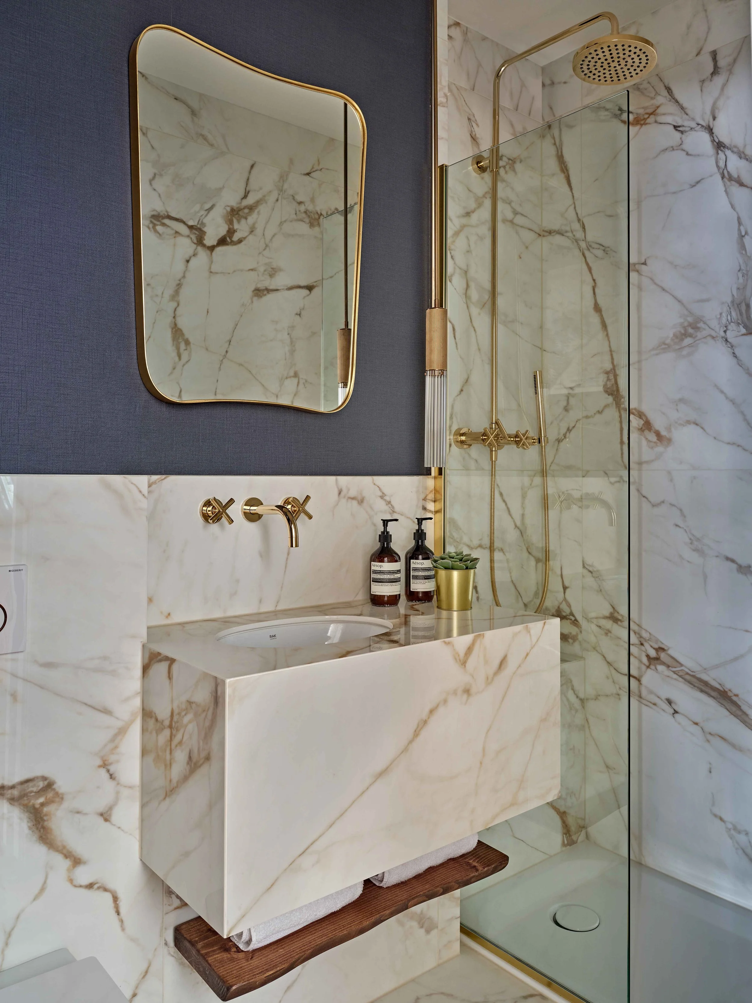 A modern bathroom featuring a marble vanity with a built-in sink, a rectangular mirror with a gold frame, and a glass-enclosed shower with a gold showerhead and fixtures. Decor includes soap dispensers and a small plant on the vanity.