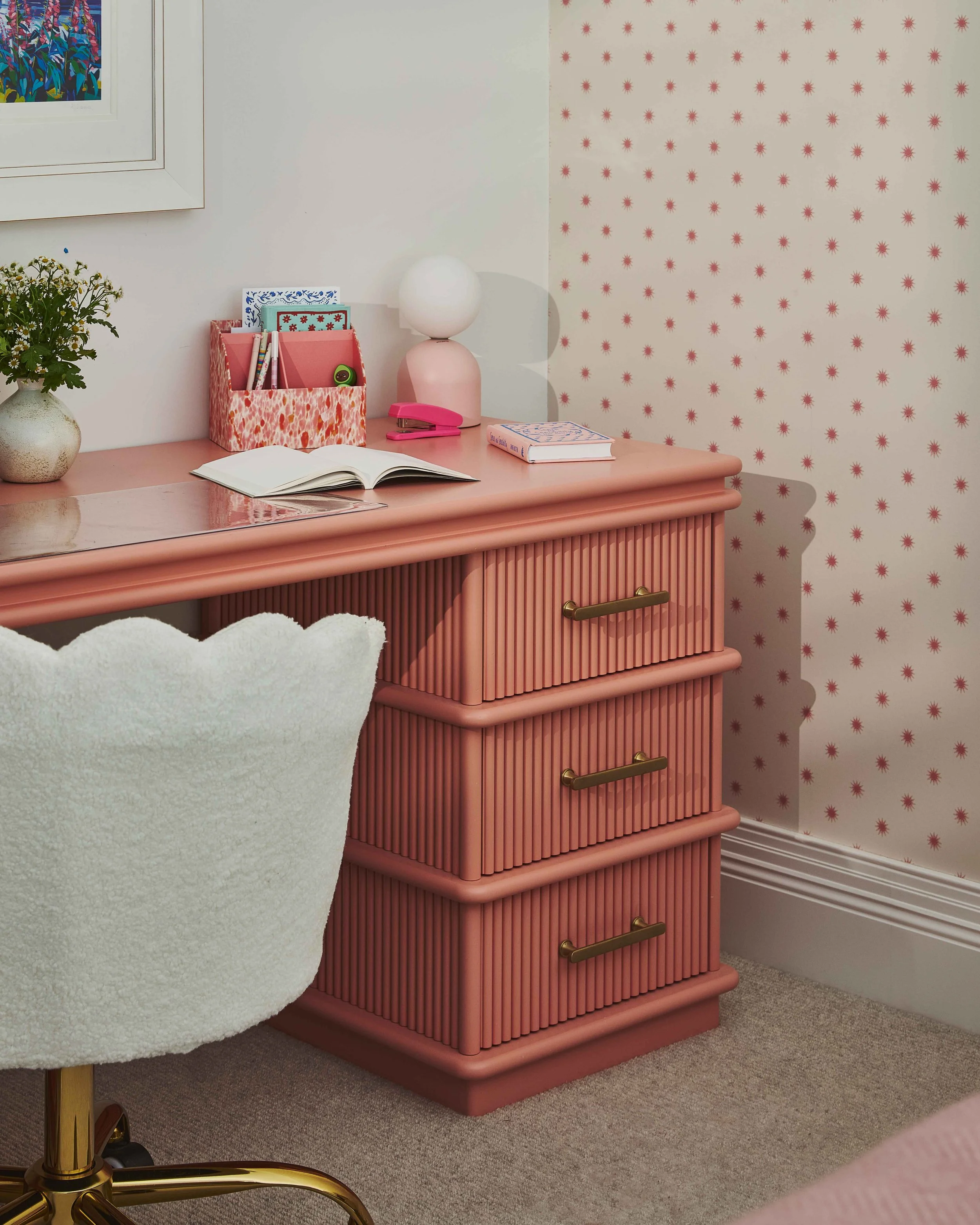 Pink dresser with gold handles against decorated wallpaper. On top of the dresser, there's an open book, a pink stapler, a pink and coral storage container with pens, a candle-shaped lamp, and a small art print. A white chair with a curvy, fluffy backrest is in front of the dresser. To the left, a white vase with green and white flowers is partially visible. The room features a framed artwork on the wall.