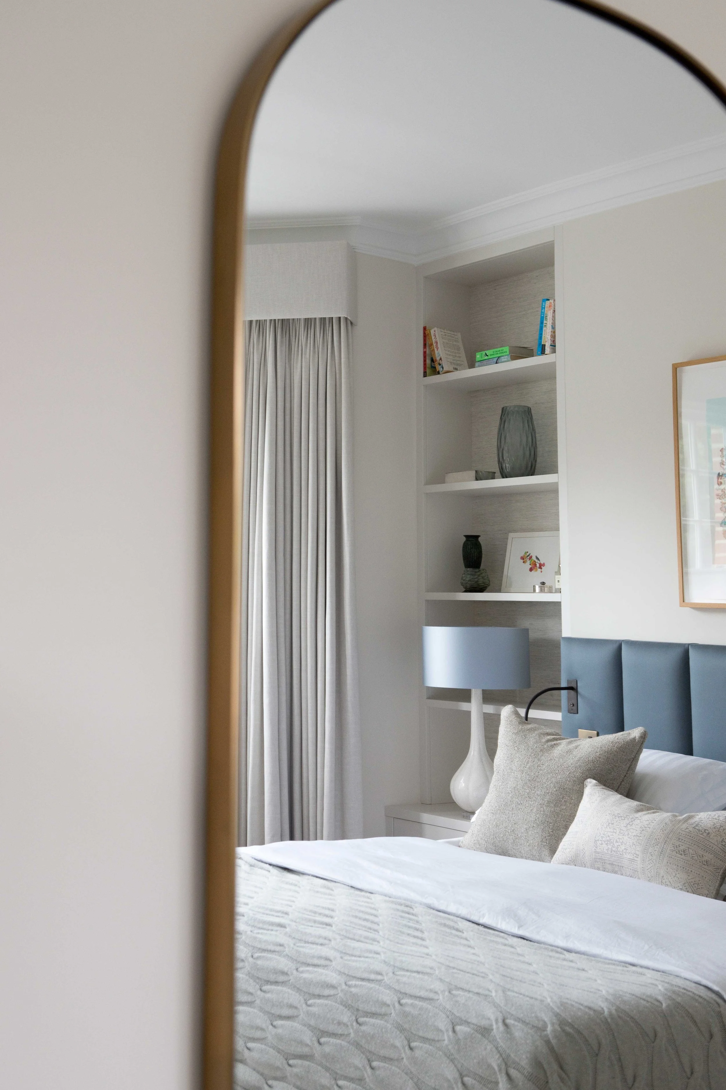 A bedroom with a corner shelf holding books and decorative vases, a grey upholstered headboard, a white side table with a white lamp, and a window with light-colored curtains, all reflected in a mirror.