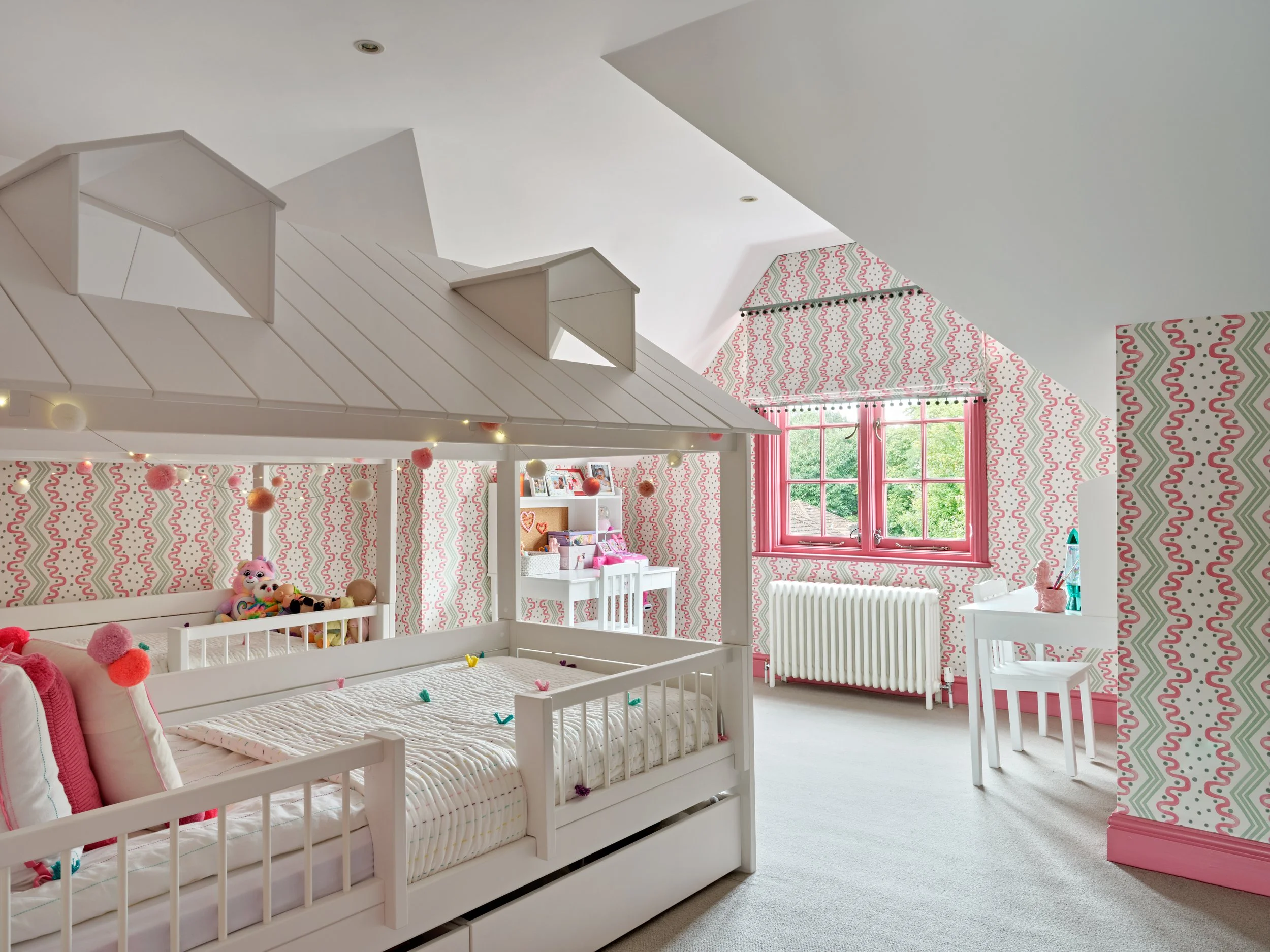 A brightly lit children's bedroom with pink and white decor, featuring a white bed with stuffed animals, pink window frames, patterned wallpaper, and a playhouse roof structure.