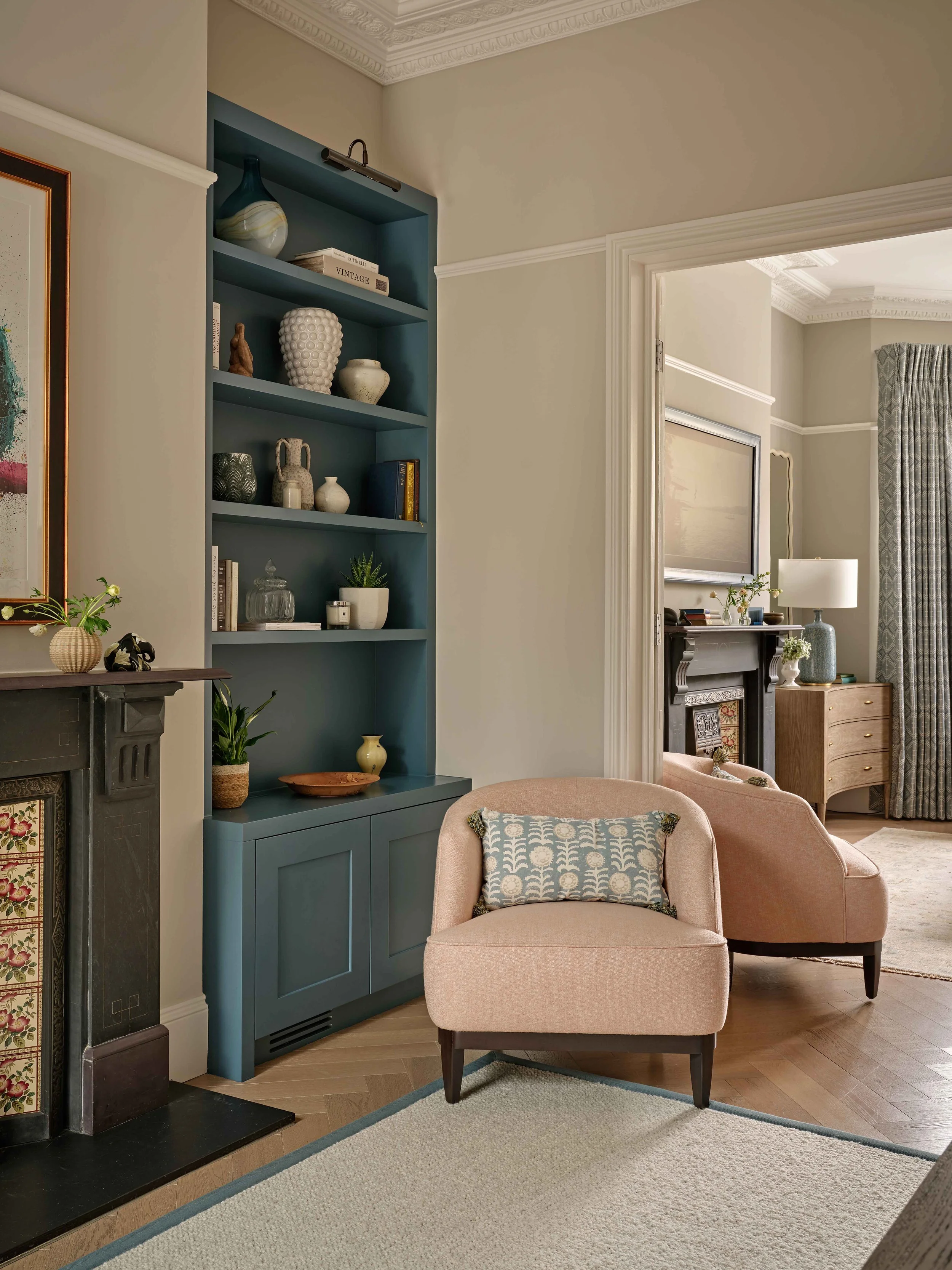 Living room corner with a pink armchair and a beige armchair, a blue bookshelf with decorative vases and books, a fireplace with a floral mosaic panel, a table with a plant, a lamp, and a cross on the wall, and a view into another room with a window,