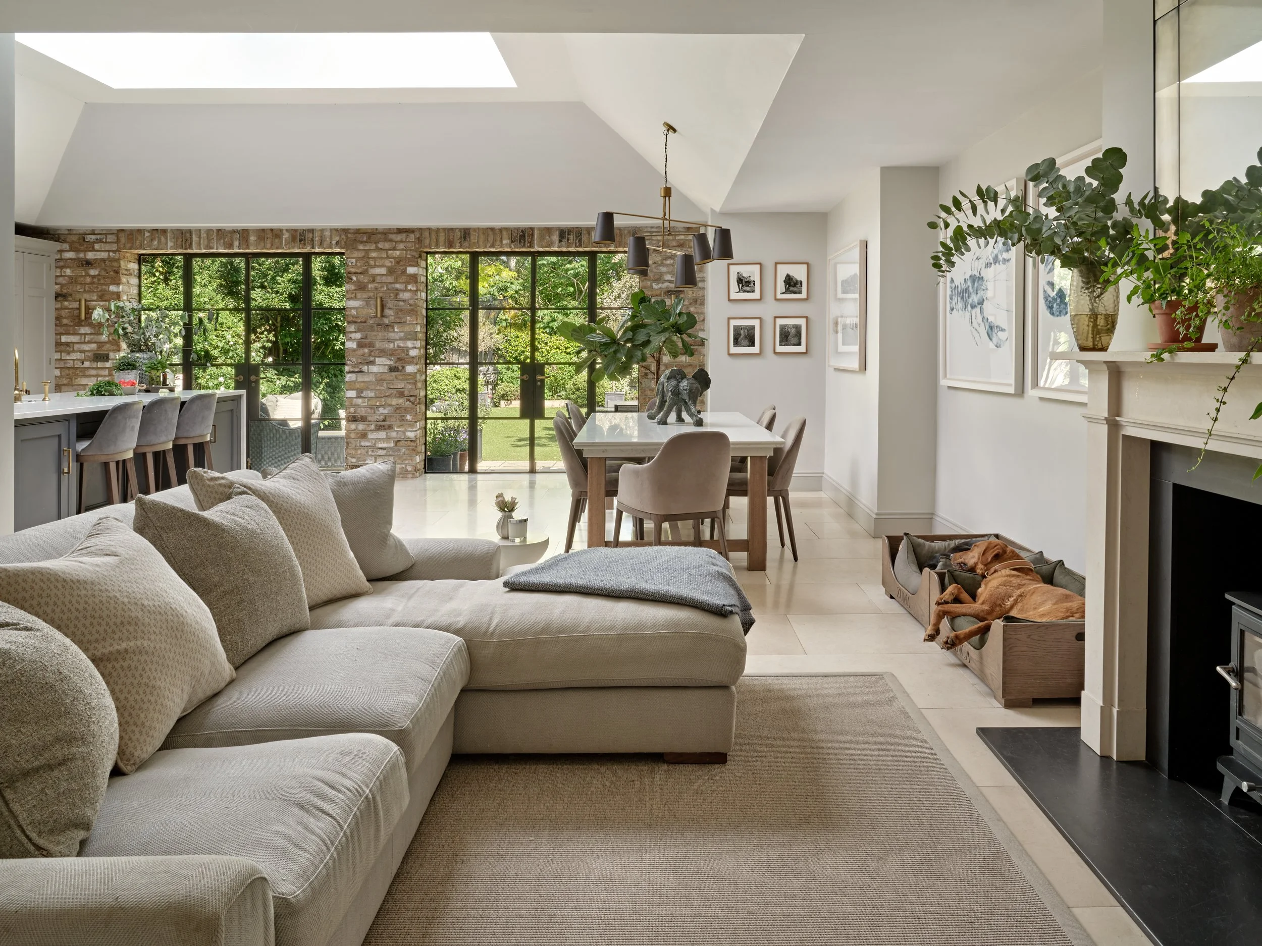 Living room with beige couch and gray pillows, hardwood floor, brick accent wall with large windows and greenery outside, dining table with beige chairs, wall art, potted plants, and a dog in a wooden bed near a fireplace.