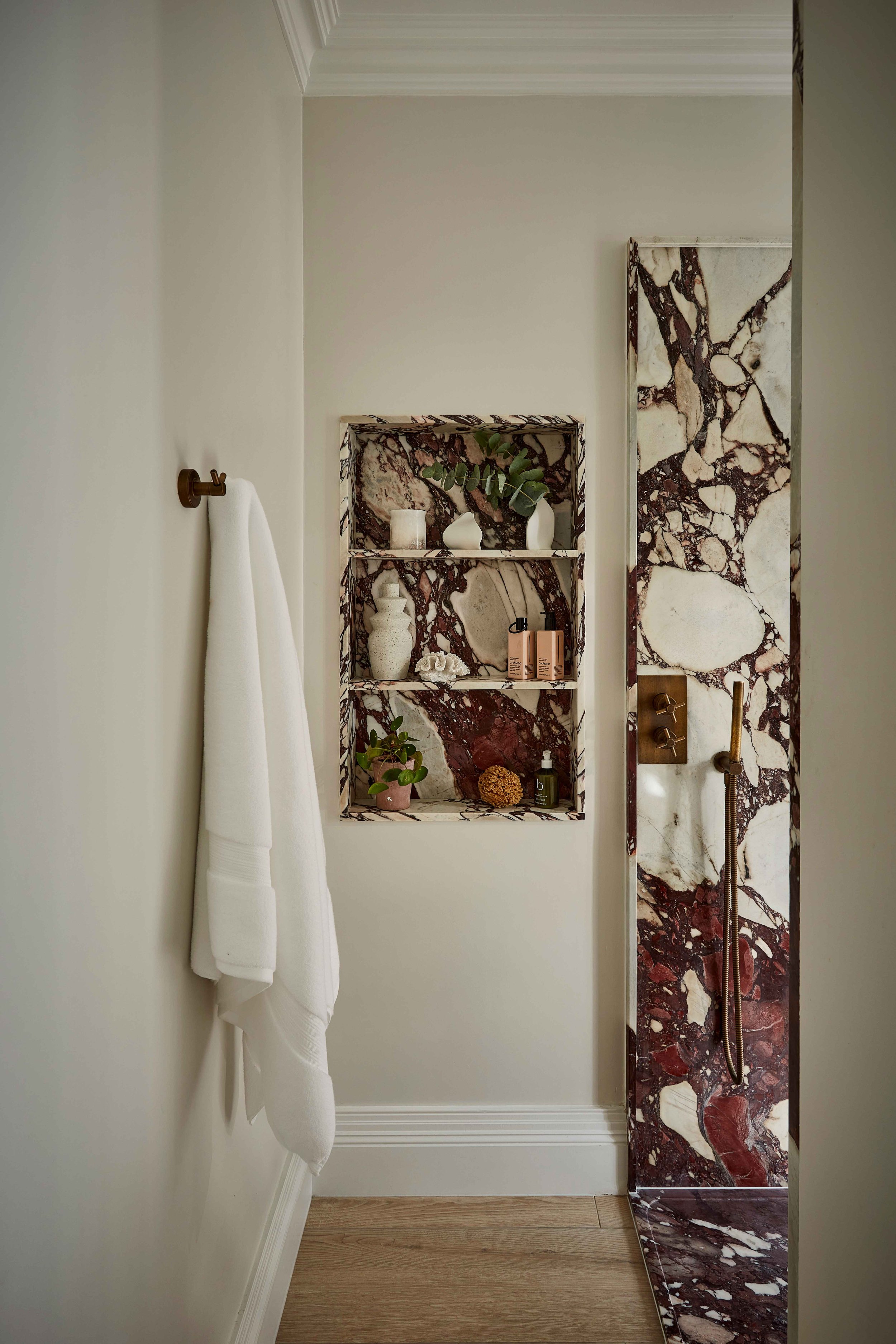 A bathroom with a white wall, a towel hanging on a brass hook, a built-in shelf with decorative items and toiletries, and a marble shower panel with a showerhead and knobs, and a wooden floor.