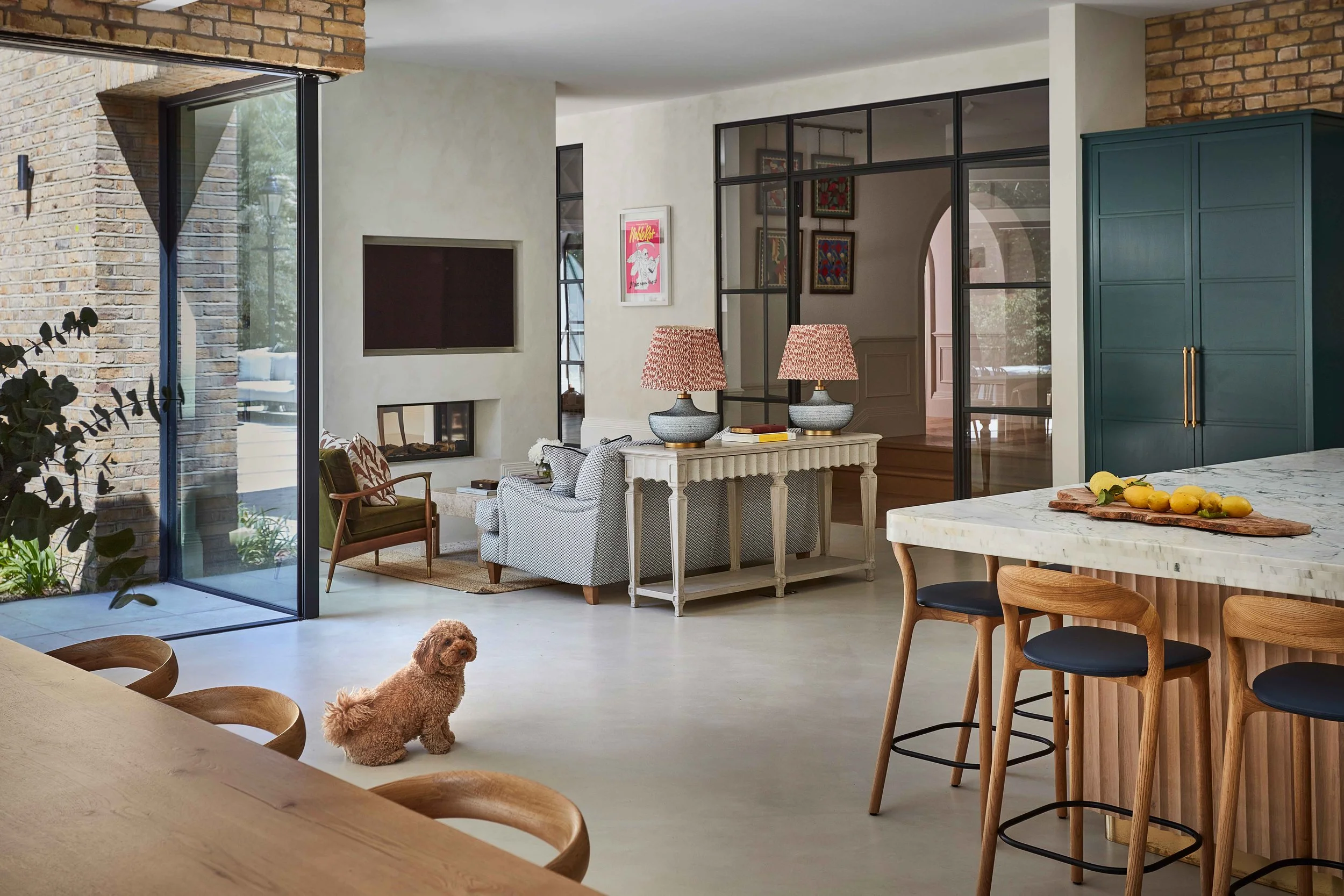 A modern living room with a brick accent wall, glass partitions, and contemporary furniture. There are two lamps with patterned shades on a white console table, and a small dog standing on the floor.