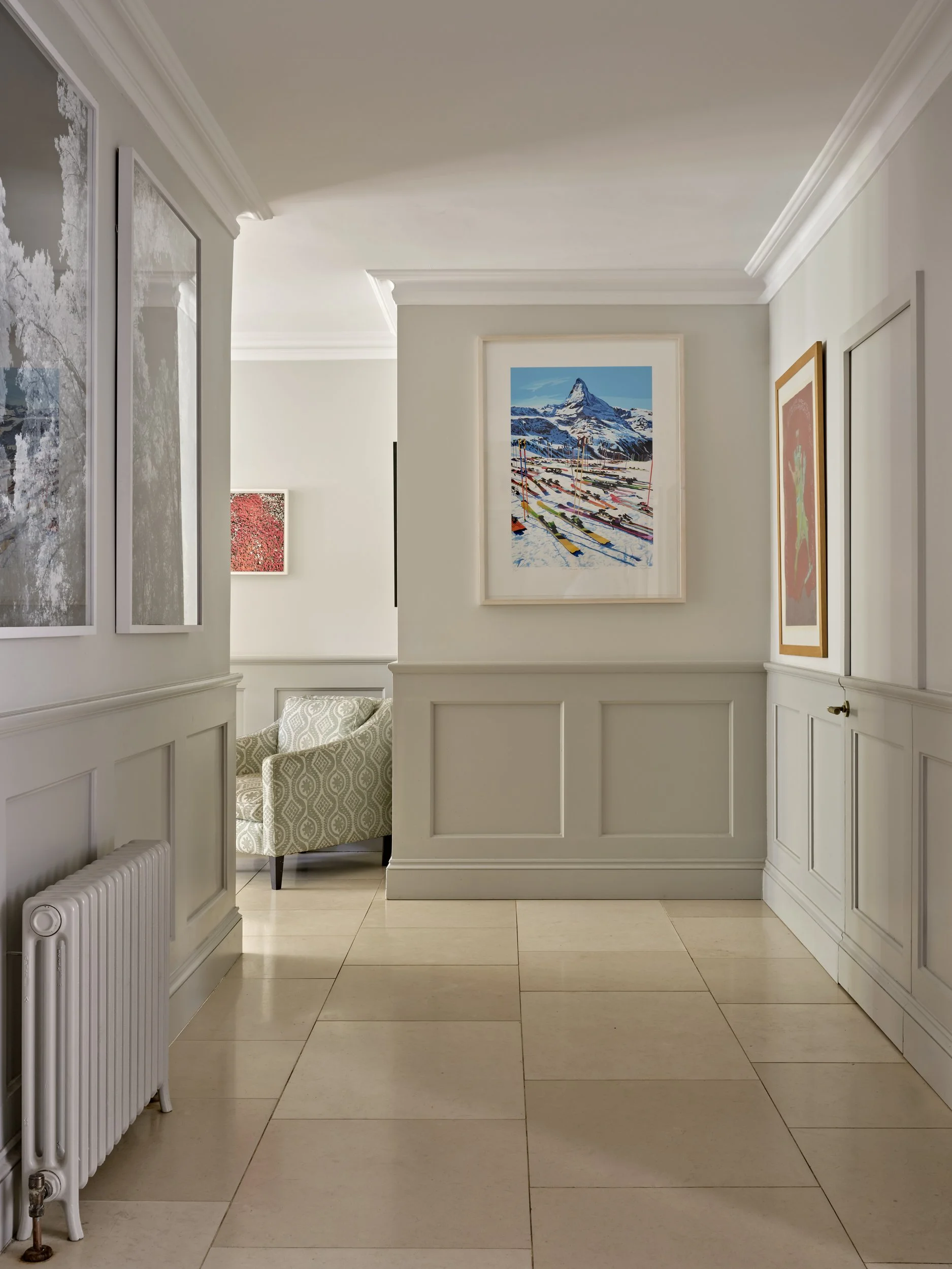 Interior hallway with framed artwork on white walls, beige tiled floor, and a patterned armchair visible in the corner.