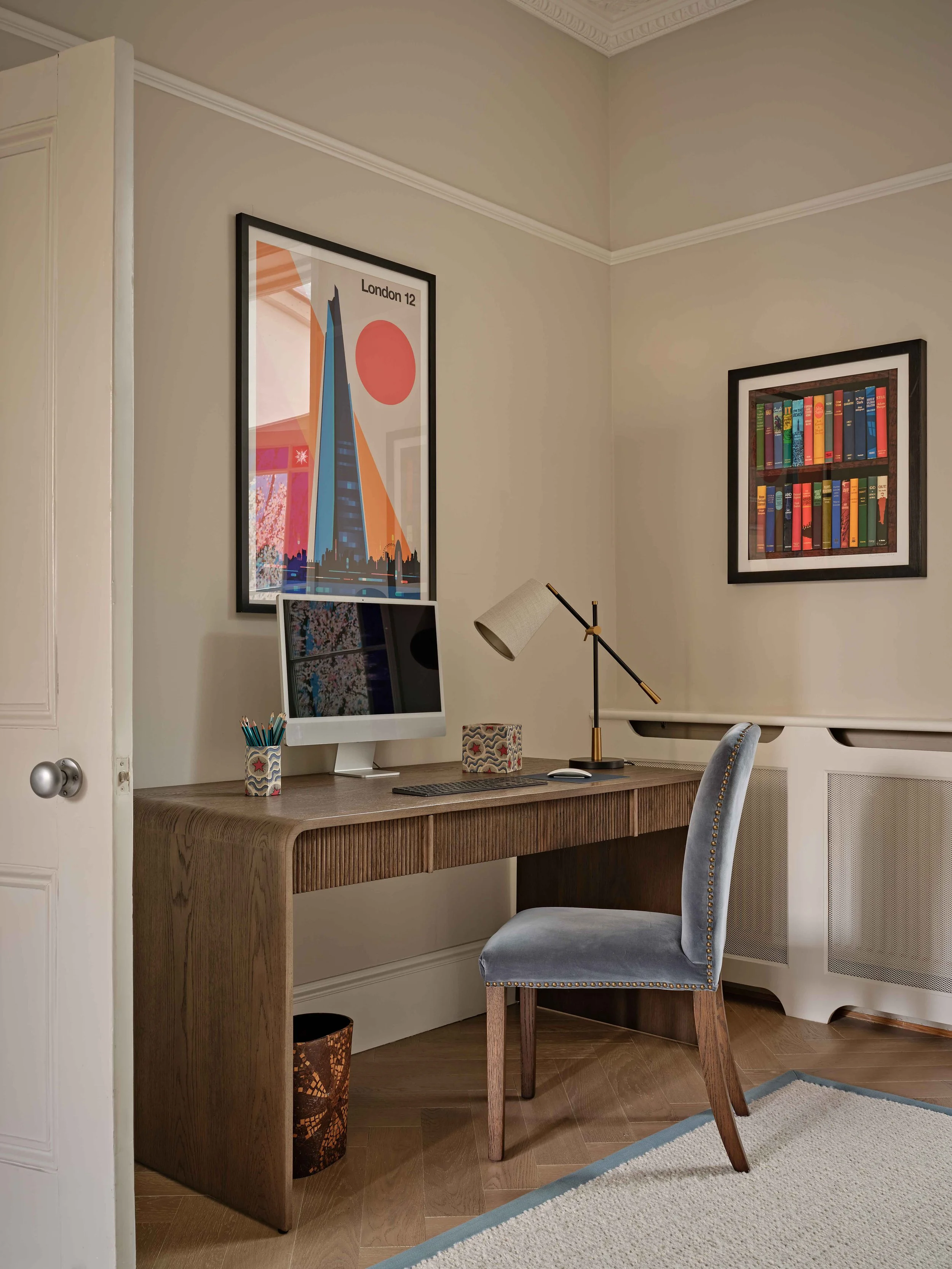 A home office corner with a wooden desk, a gray upholstered chair, a modern desk lamp, a computer monitor, a pen holder, framed art posters on the wall, and a beige rug on wooden flooring.