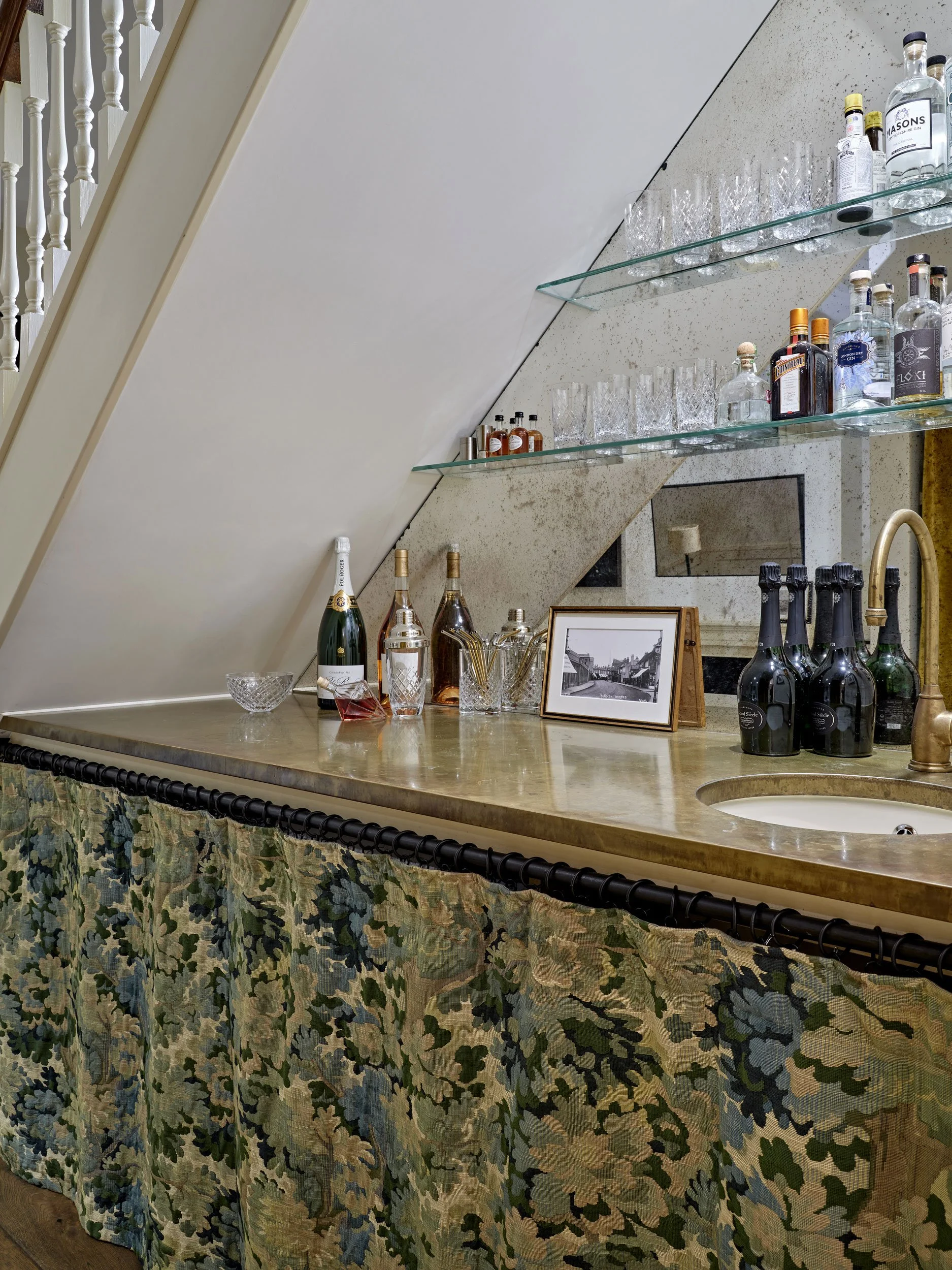 A bar area with a yellow marble countertop, decorated with a camo patterned fabric skirt. On the counter are wine bottles, glasses, a framed black-and-white photo, and some drink mixers. A nearby wall has shelves with assorted liquor bottles and glas