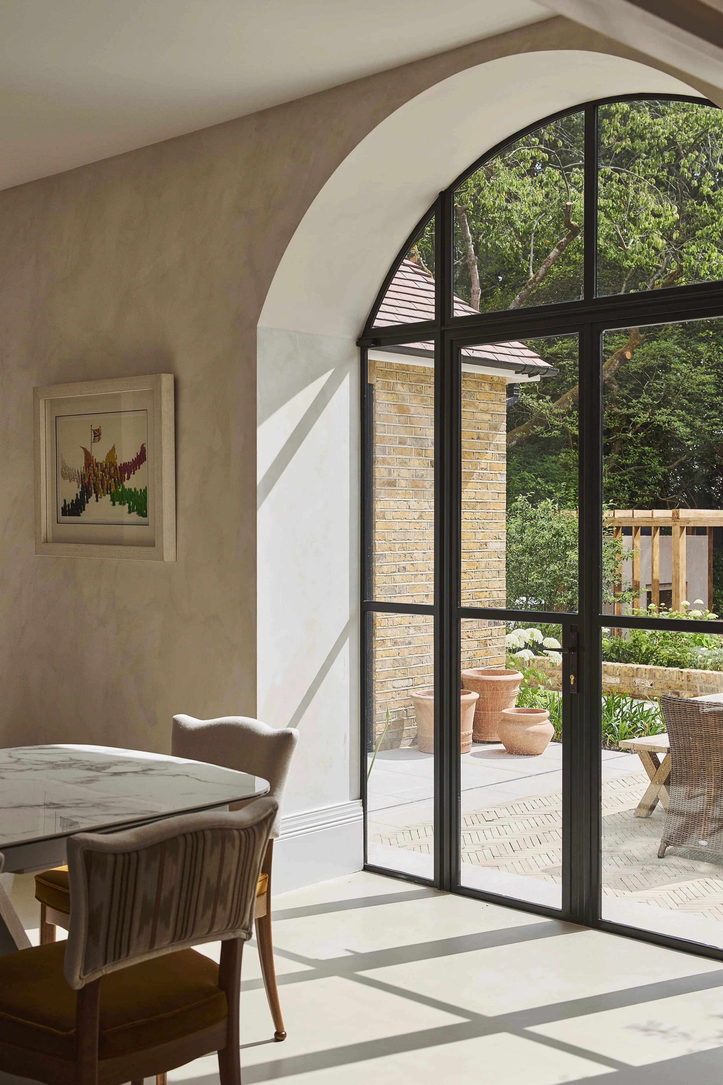 Sunlit interior of a room with a round table and striped chair, large arched window overlooking a patio garden with potted plants and trees.