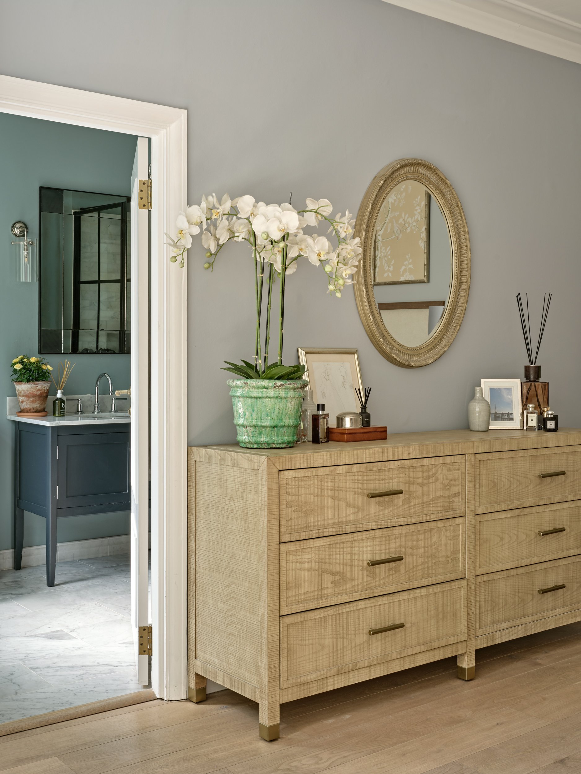 A wooden dresser with gold handles and a tall green potted orchid on top, placed against a wall with a gold oval mirror and framed artwork, in a room with hardwood floors.