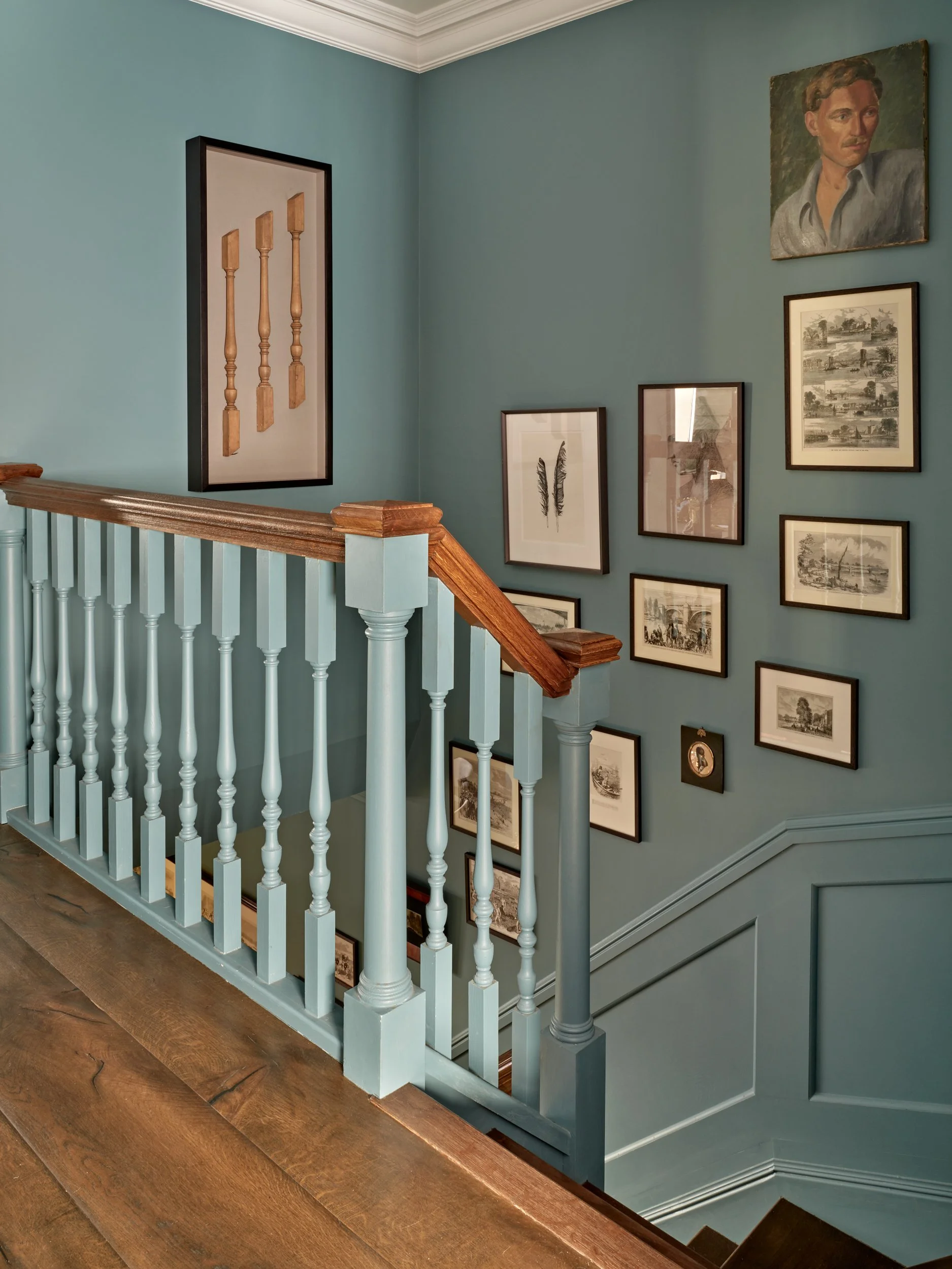 A staircase with white spindles and a wooden handrail leading down to a lower floor, with a wall decorated with framed artwork including a portrait painting and sketches, on a blue-green wall.