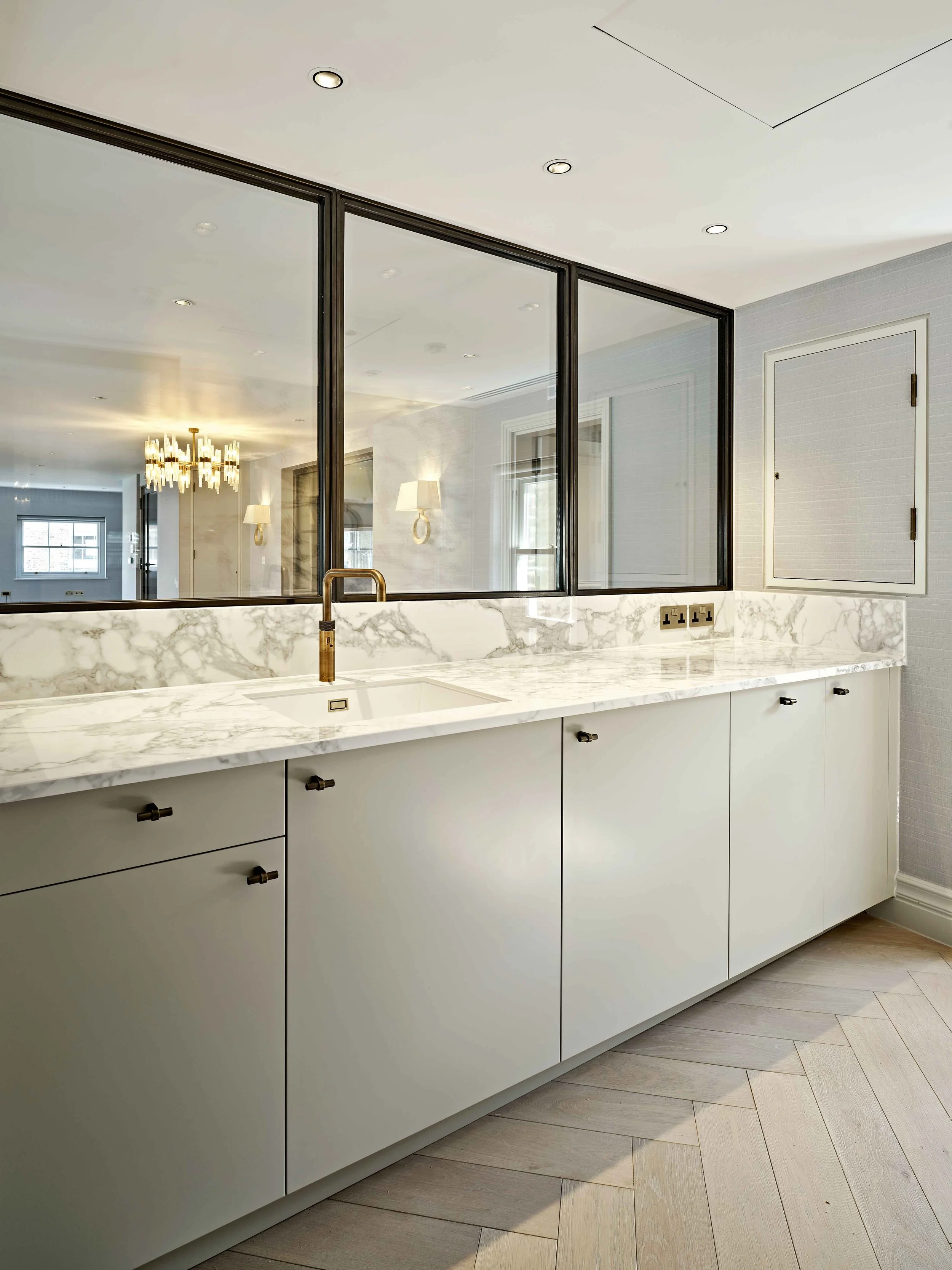 Modern kitchen with white cabinetry, marble countertop, black hardware, and a large mirror reflecting a chandelier and window.