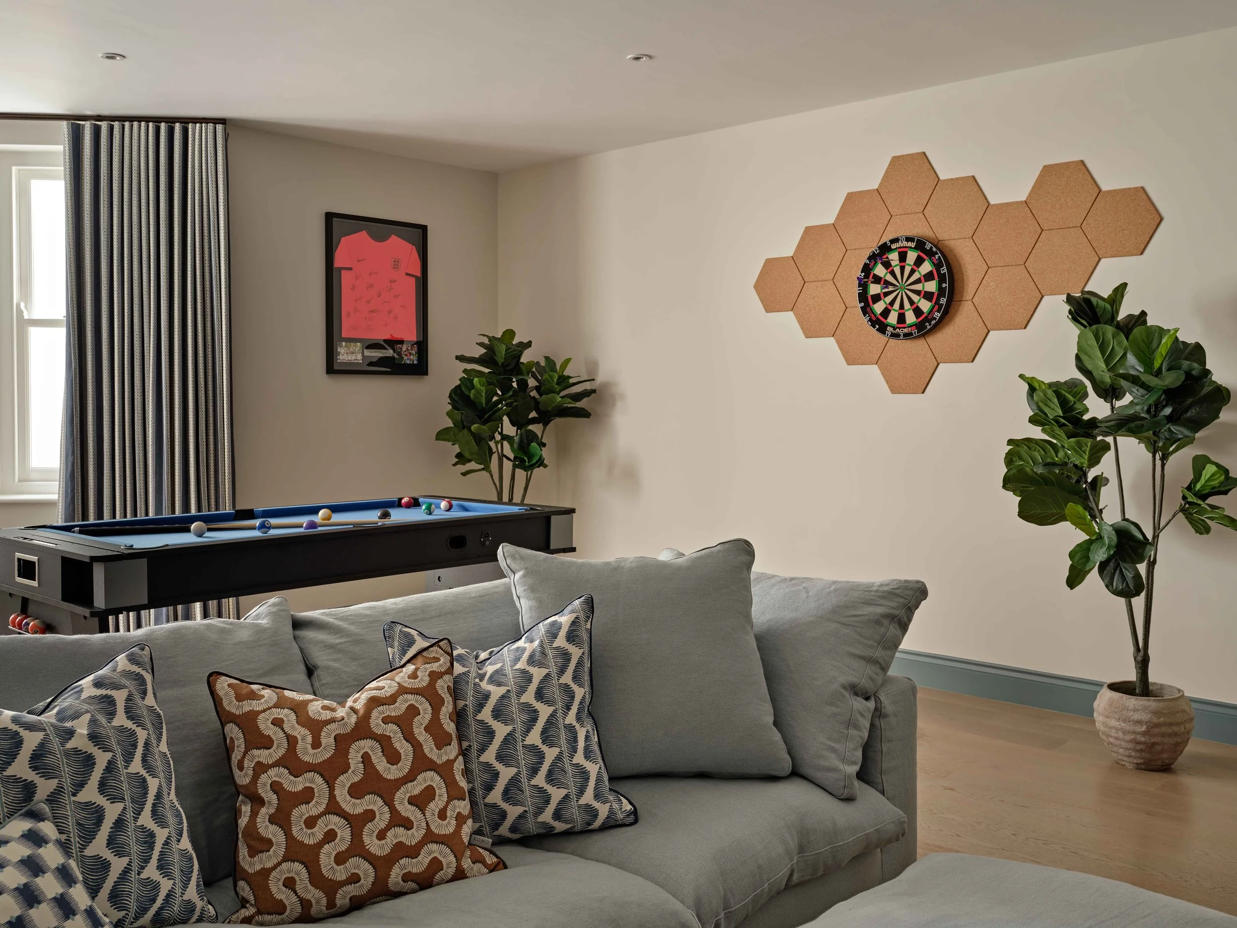 Living room with a pool table, framed sports jersey on wall, dartboard on wall surrounded by cork hexagon panels, houseplants, and sofa with patterned pillows.