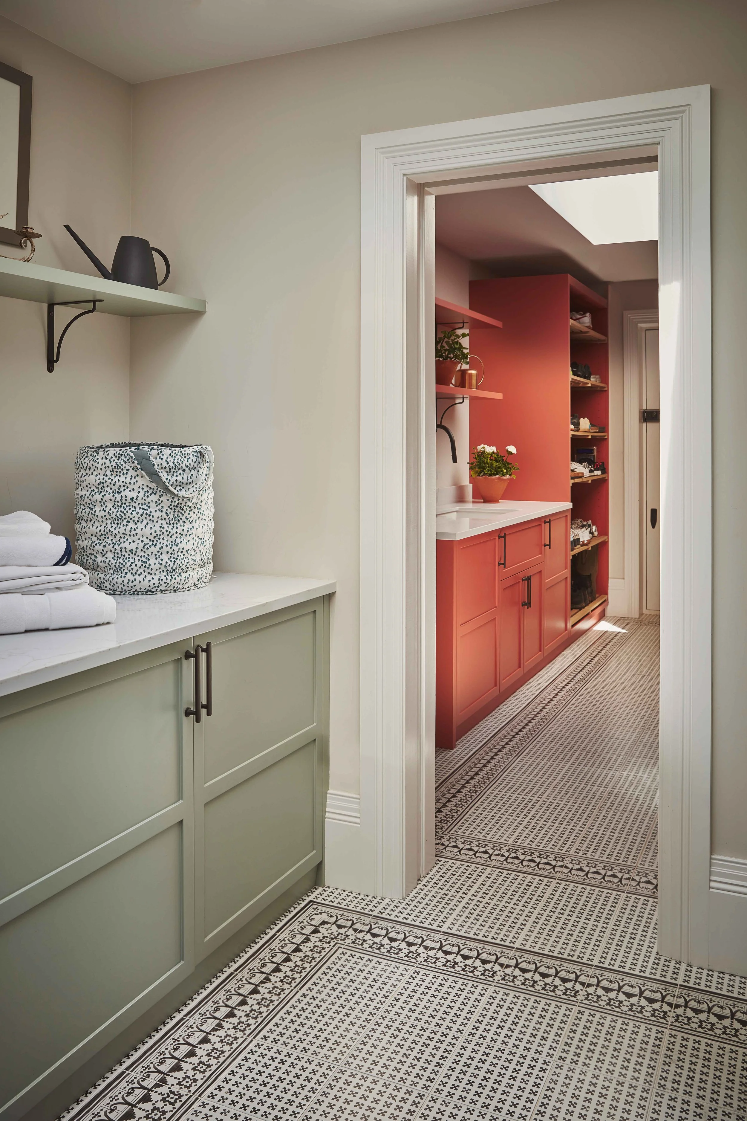Interior view showing a doorway, a green cabinet with folded towels, a basket, and a countertop with potted plants, leading to a room with red cabinetry, a patterned rug, and natural light.