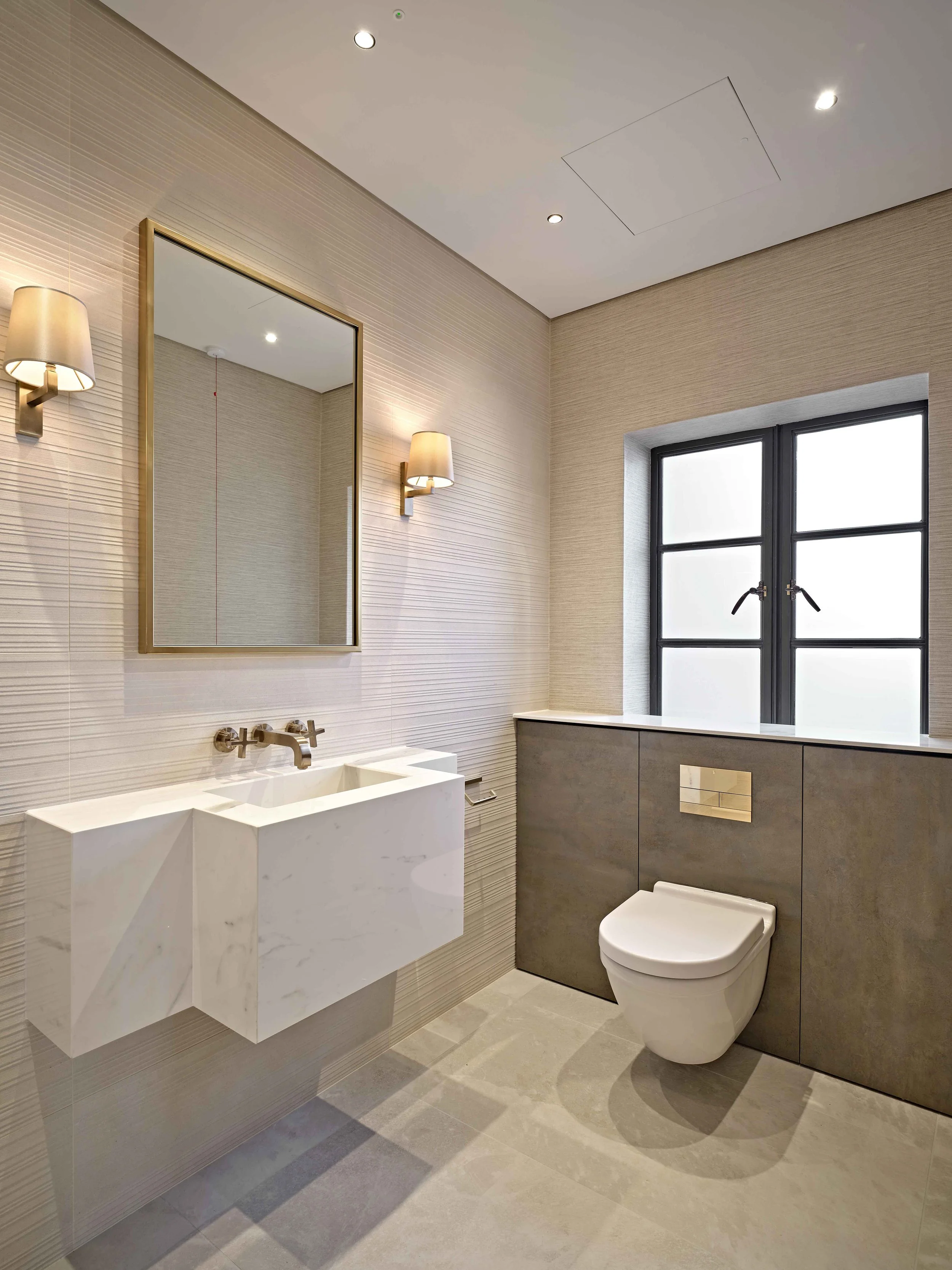 Modern bathroom with a wall-mounted white marble sink, a large mirror framed in gold, wall-mounted beige and gold lighting fixtures, a frosted window, and a sleek white toilet. The walls have textured beige tiles and the floor has large beige tiles.