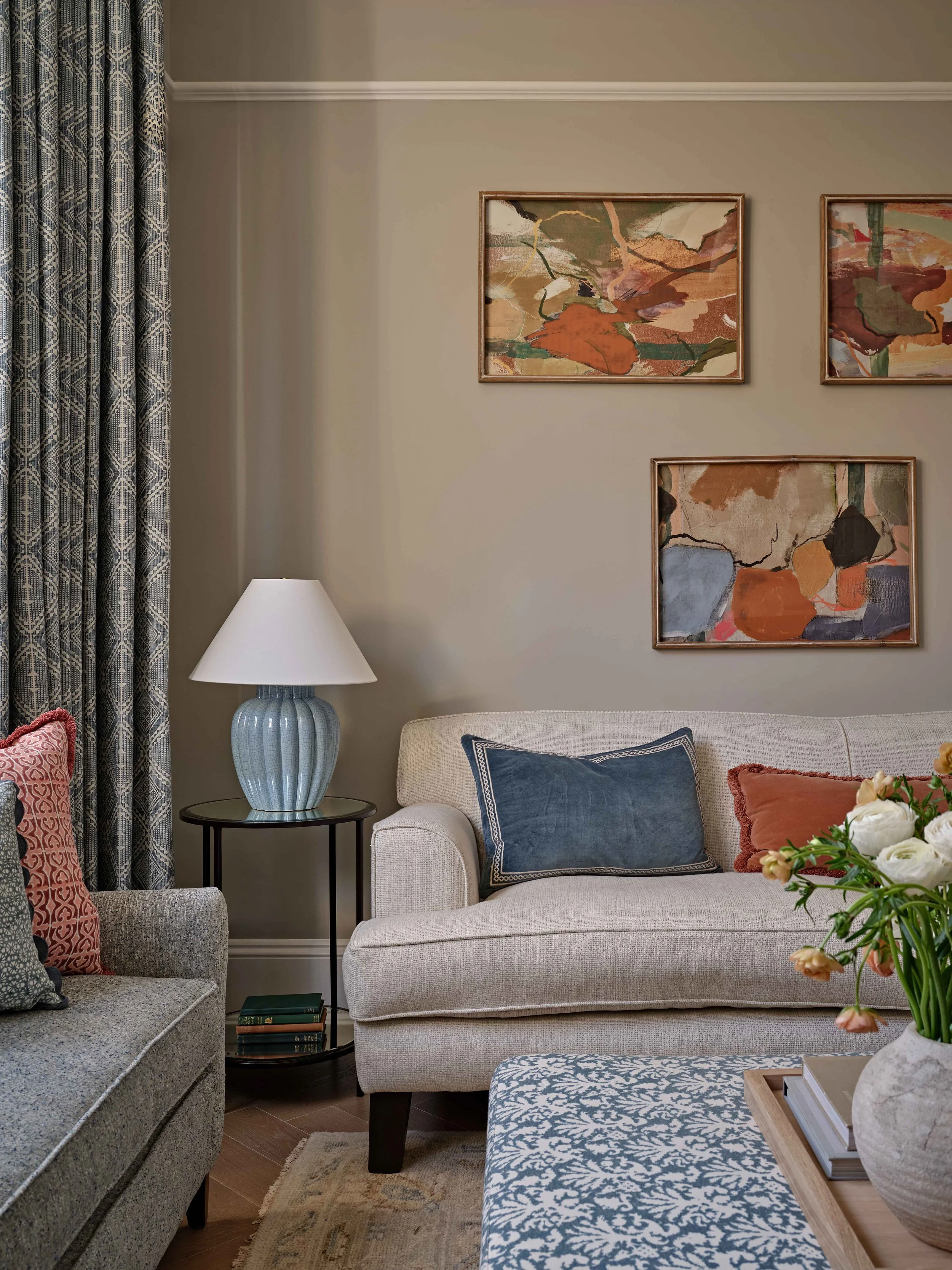 Living room with a beige sofa, decorative pillows, a side table with books, a table lamp, wall art, and floral arrangement.