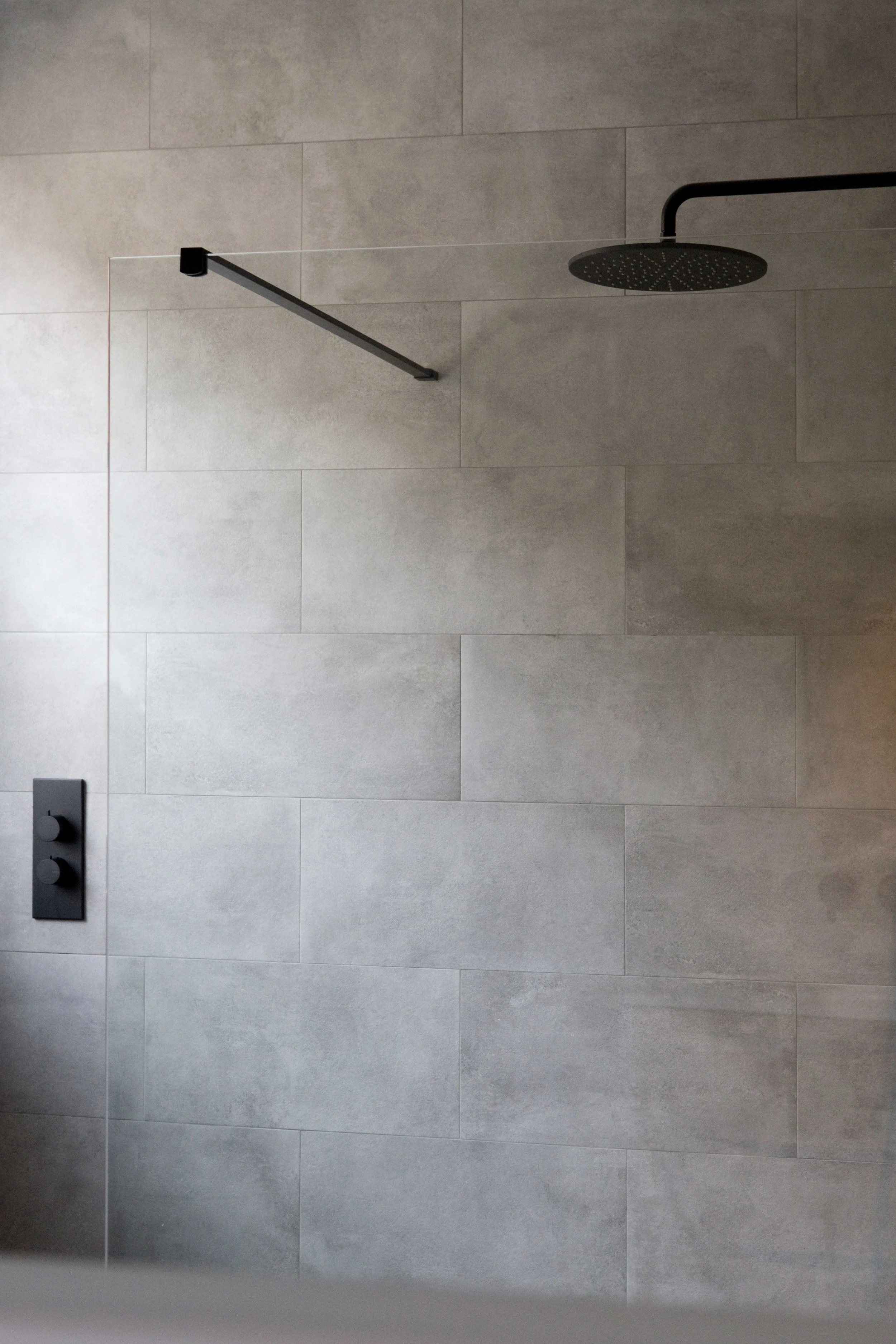 A modern walk-in shower with a large, circular, black rainfall showerhead on a gray tiled wall. The shower has a transparent glass divider and a black control panel with two knobs.