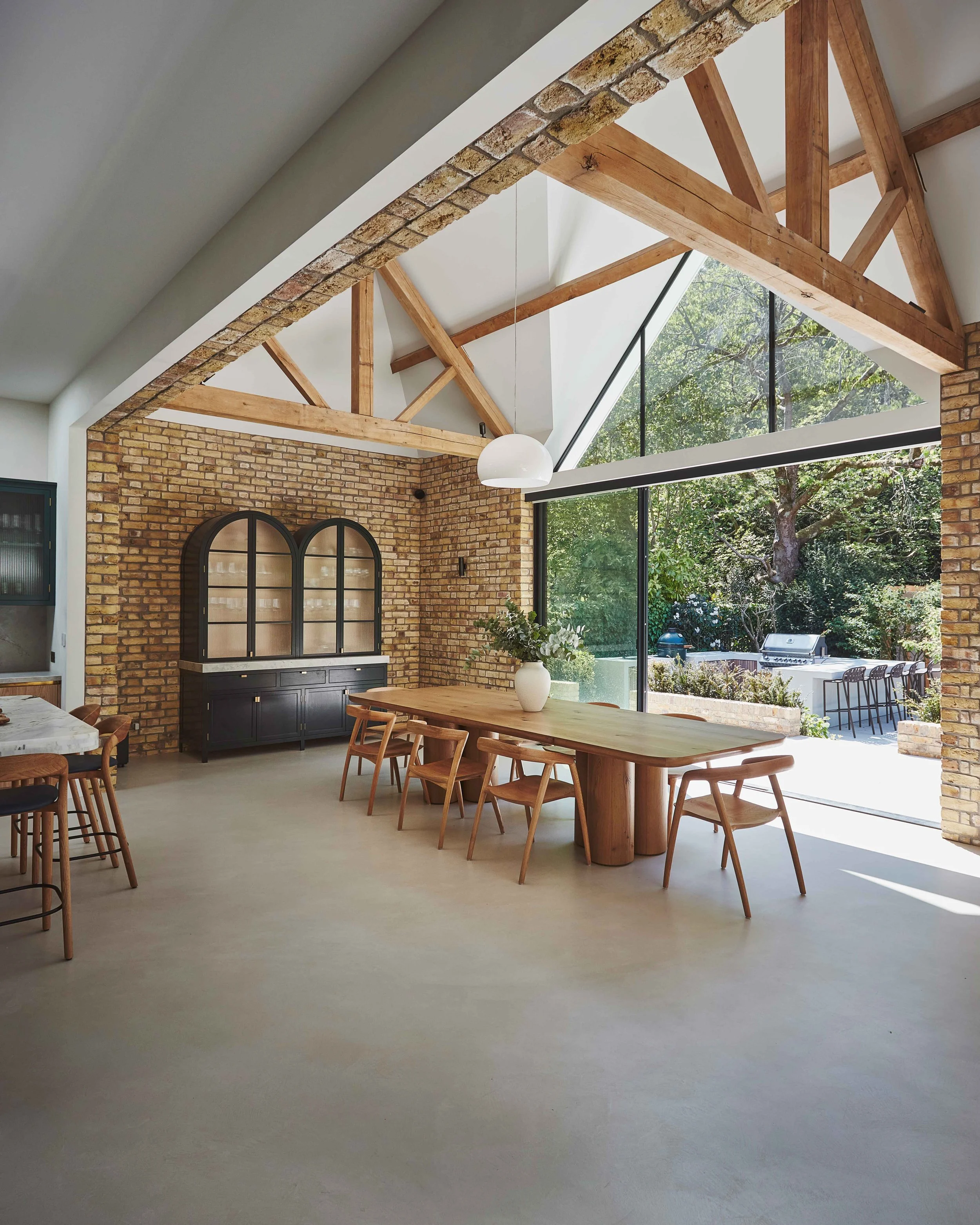 Modern dining area with wood and brick accents, large windows, wooden dining table, chairs, and a view of a backyard with outdoor seating and a grill.