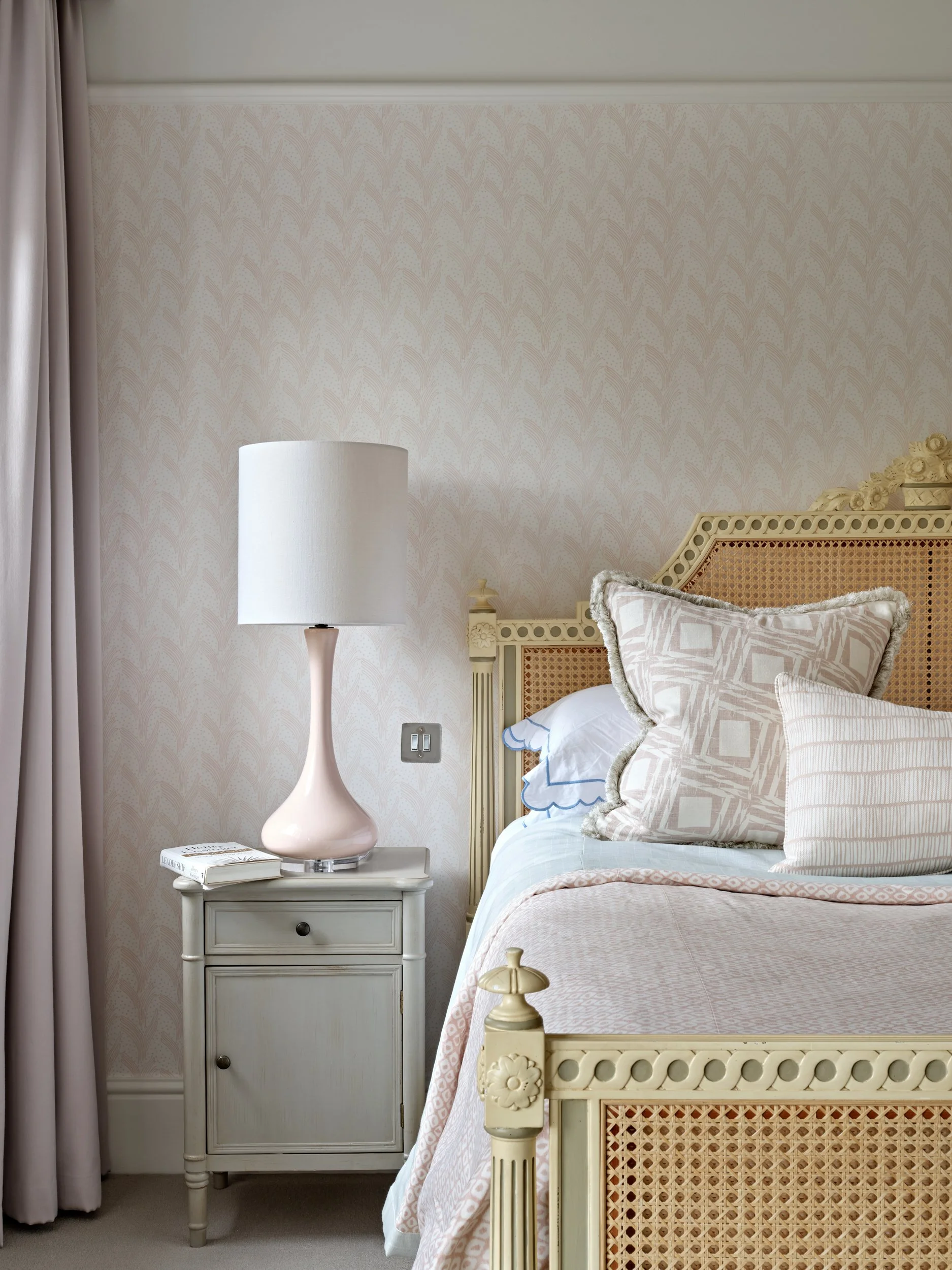 A bedroom with a white nightstand, a tall white lamp, a bed with a cream-colored headboard, pillows, and a pink-patterned blanket. Light-colored walls with subtle wallpaper, beige carpet, and cream curtains.