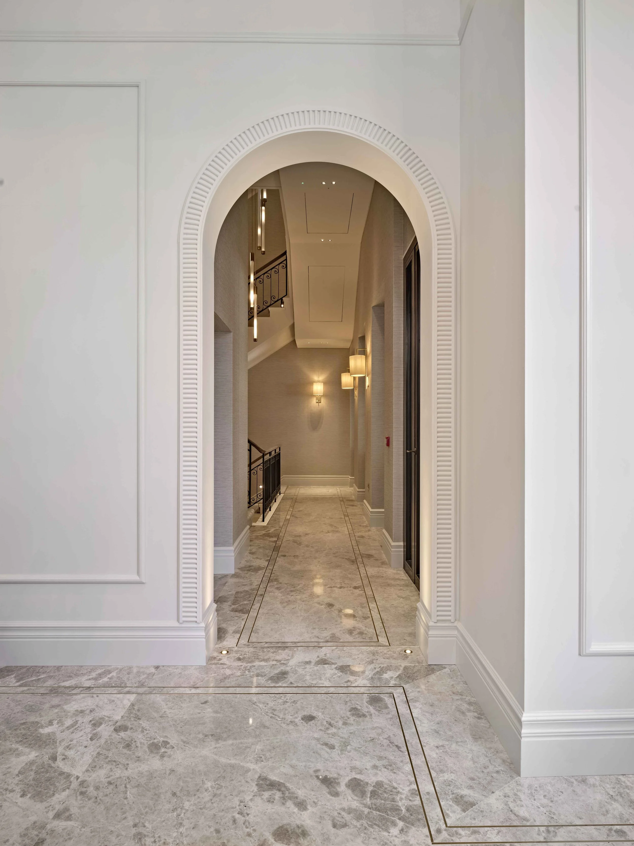 Elegant hallway with marble flooring, white walls with decorative molding, and an arched doorway leading to a staircase with modern lighting fixtures.