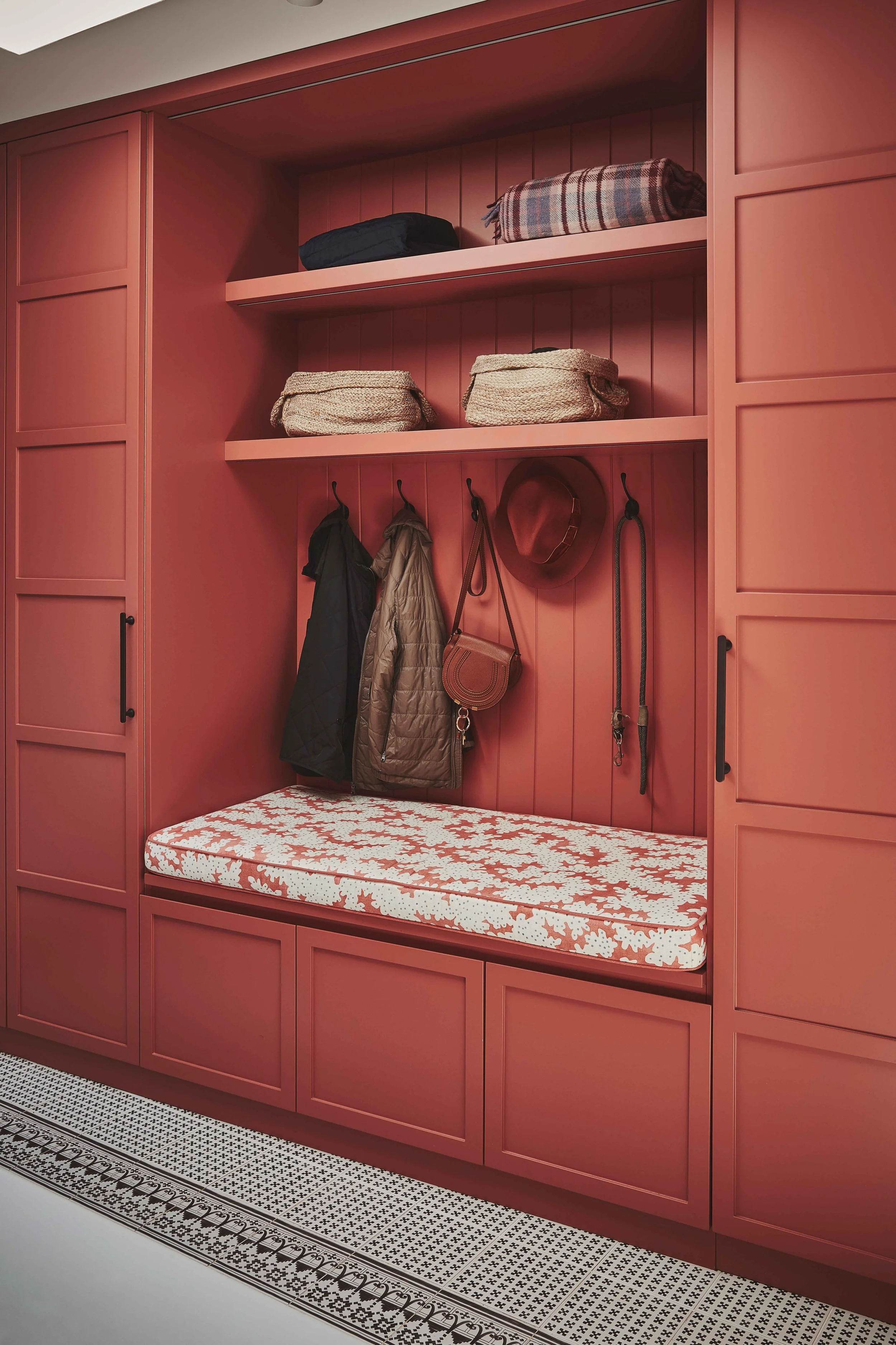 A built-in pink wardrobe with shelving and hooks. The upper shelves hold folded blankets, and the hooks below hold coats, a hat, and bags. There is a cushioned seating area with a patterned cover beneath the hooks.