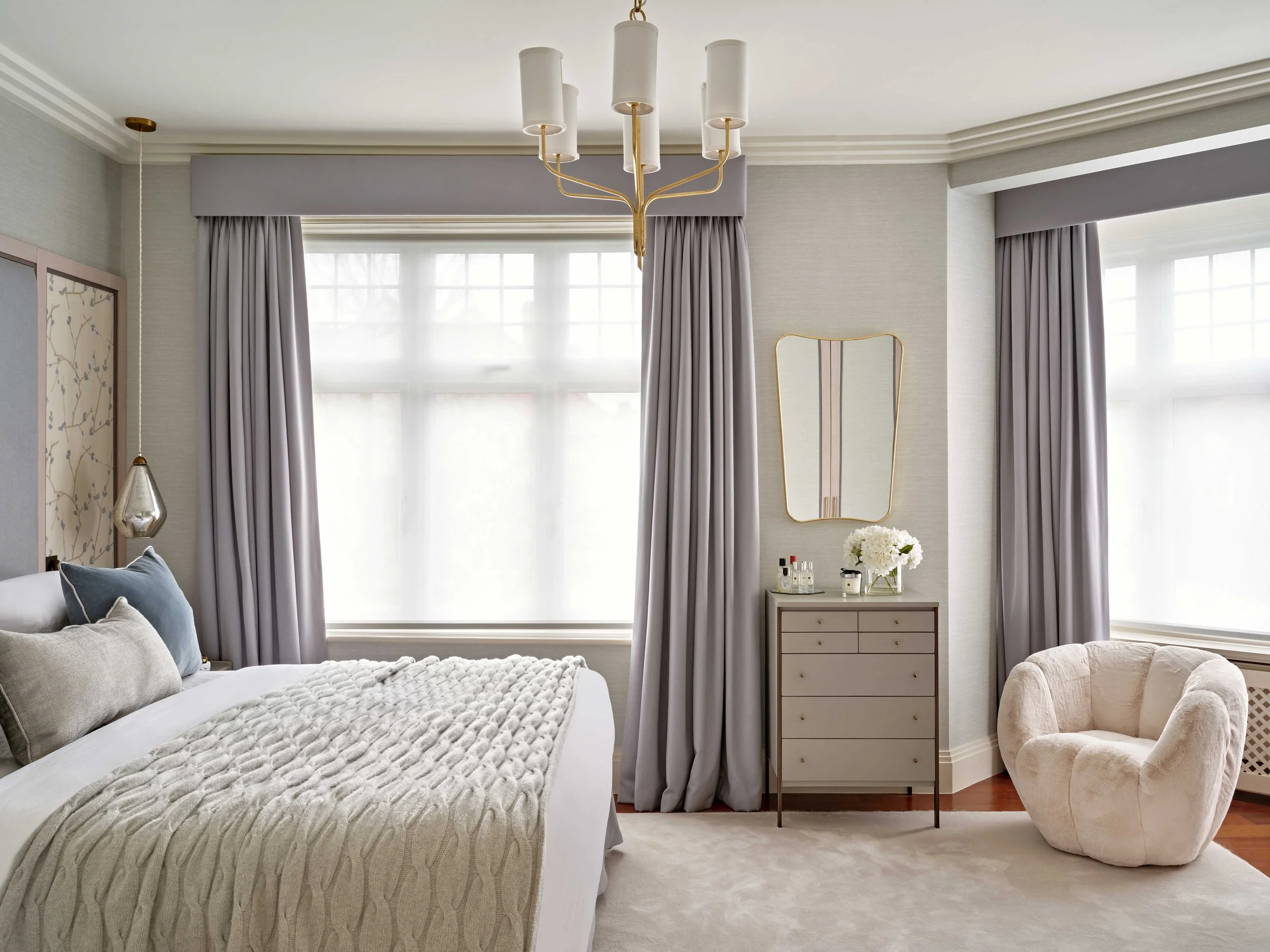 A bedroom with large windows covered by light gray curtains, a beige armchair, a white dresser with flowers, a gold mirror, and a modern chandelier hanging from the ceiling.