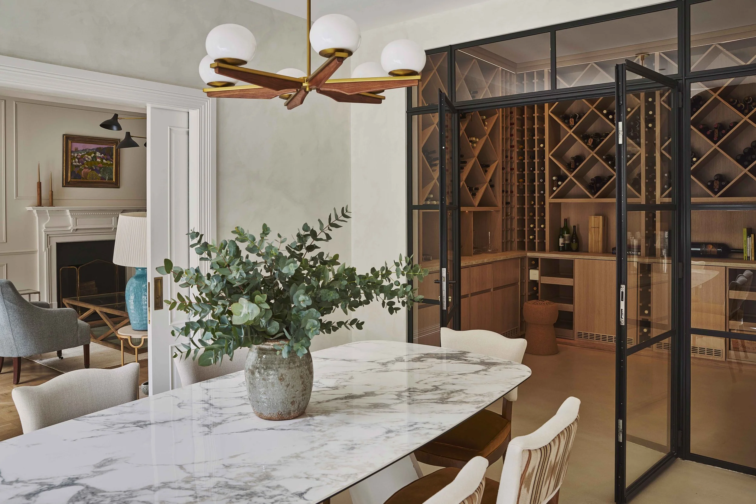 Living room with a fireplace, gray armchair, side table with a lamp, and a colorful painting on the wall. In the foreground, a marble dining table with a potted plant, and a wine cellar with wooden shelves behind a glass door.