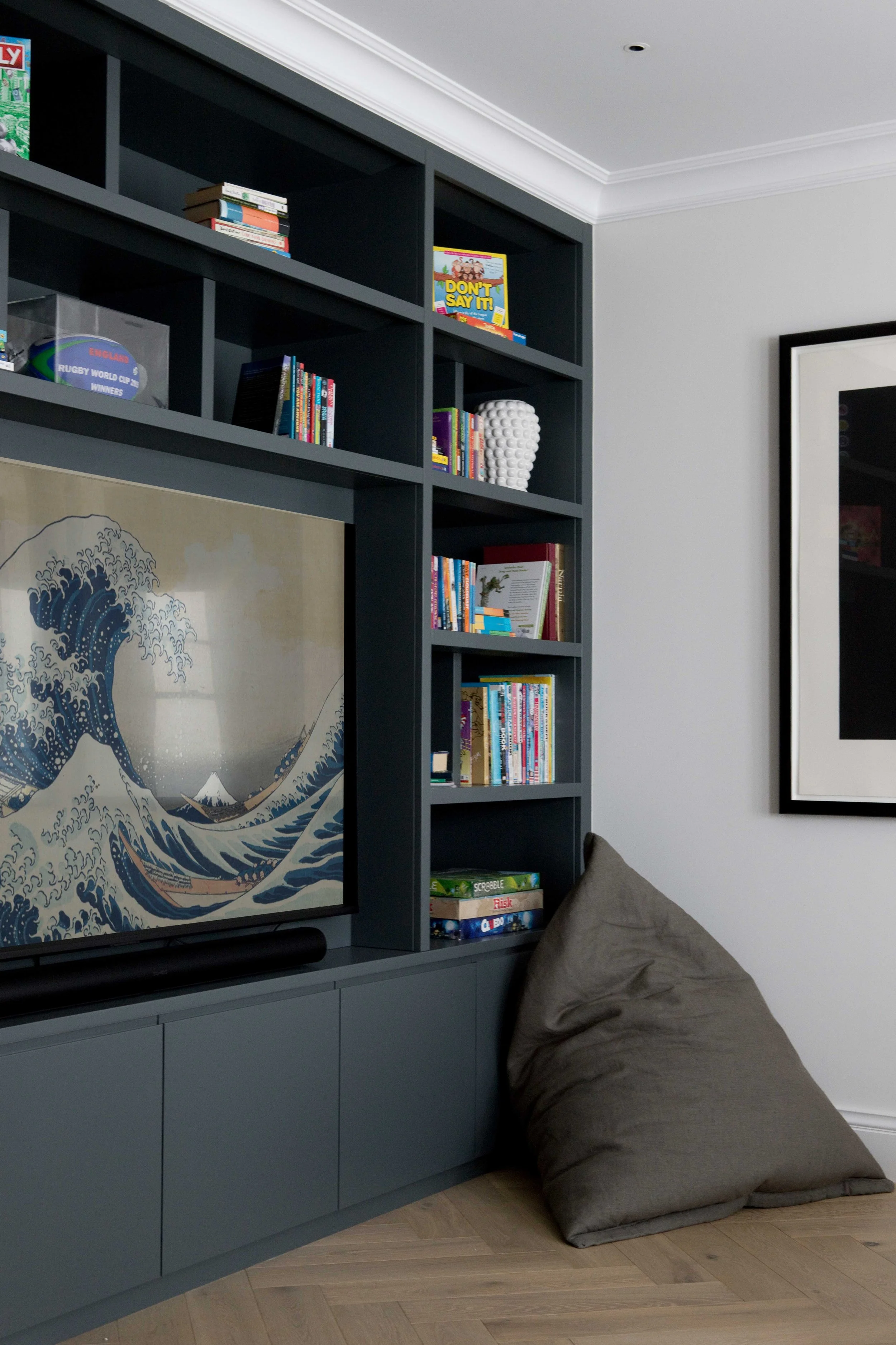 A framed artwork of a famous Japanese wave print in blue tones, a dark blue bookshelf with books and decorative objects, a gray beanbag against a gray wall, and a light wood floor in a modern living space.