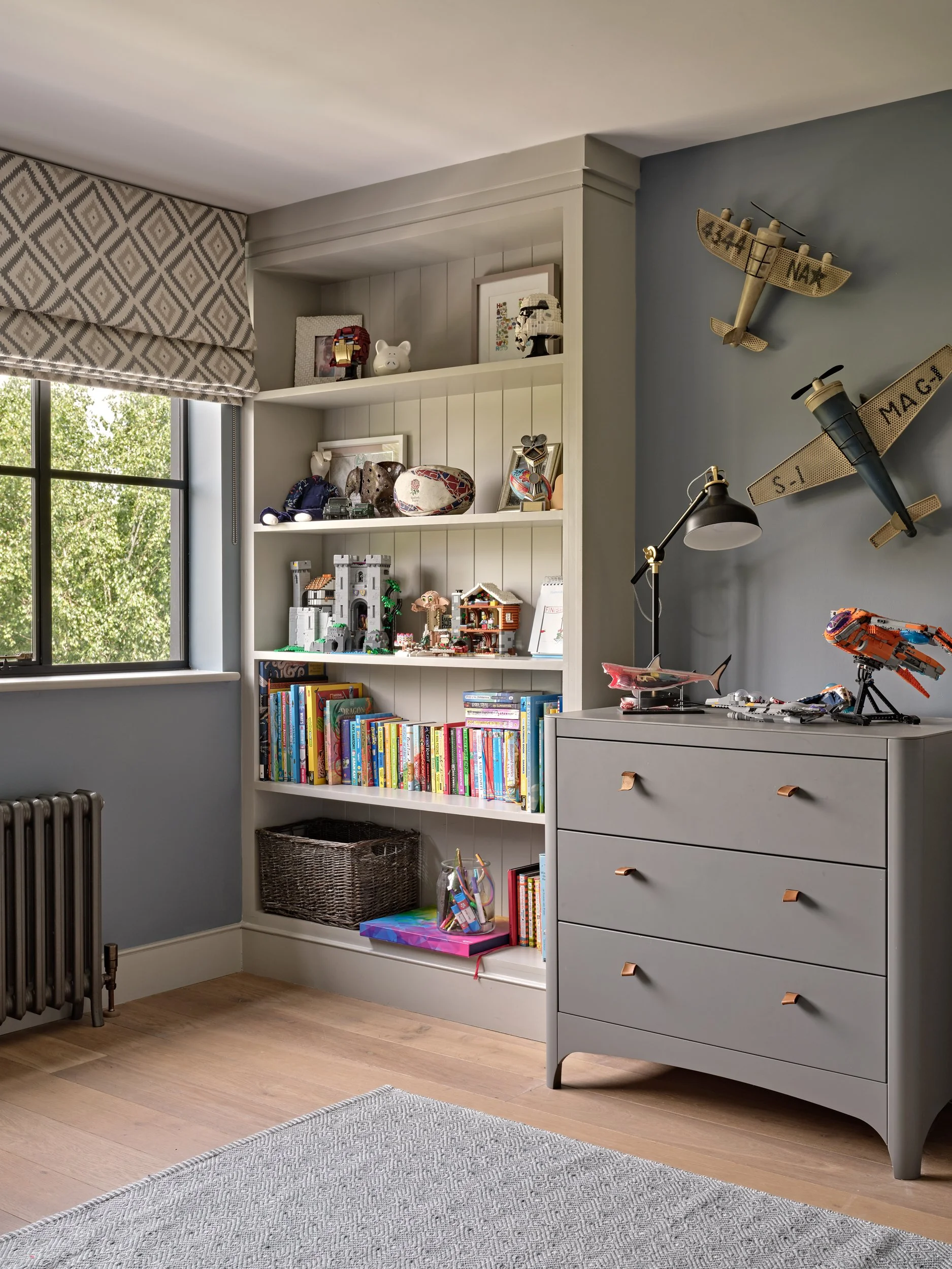 Interior of a child's bedroom with white bookshelf filled with books, toys, and framed pictures, gray dresser with Lego models, decorative plane on the wall, window with patterned Roman shades, wooden flooring, and a gray rug.