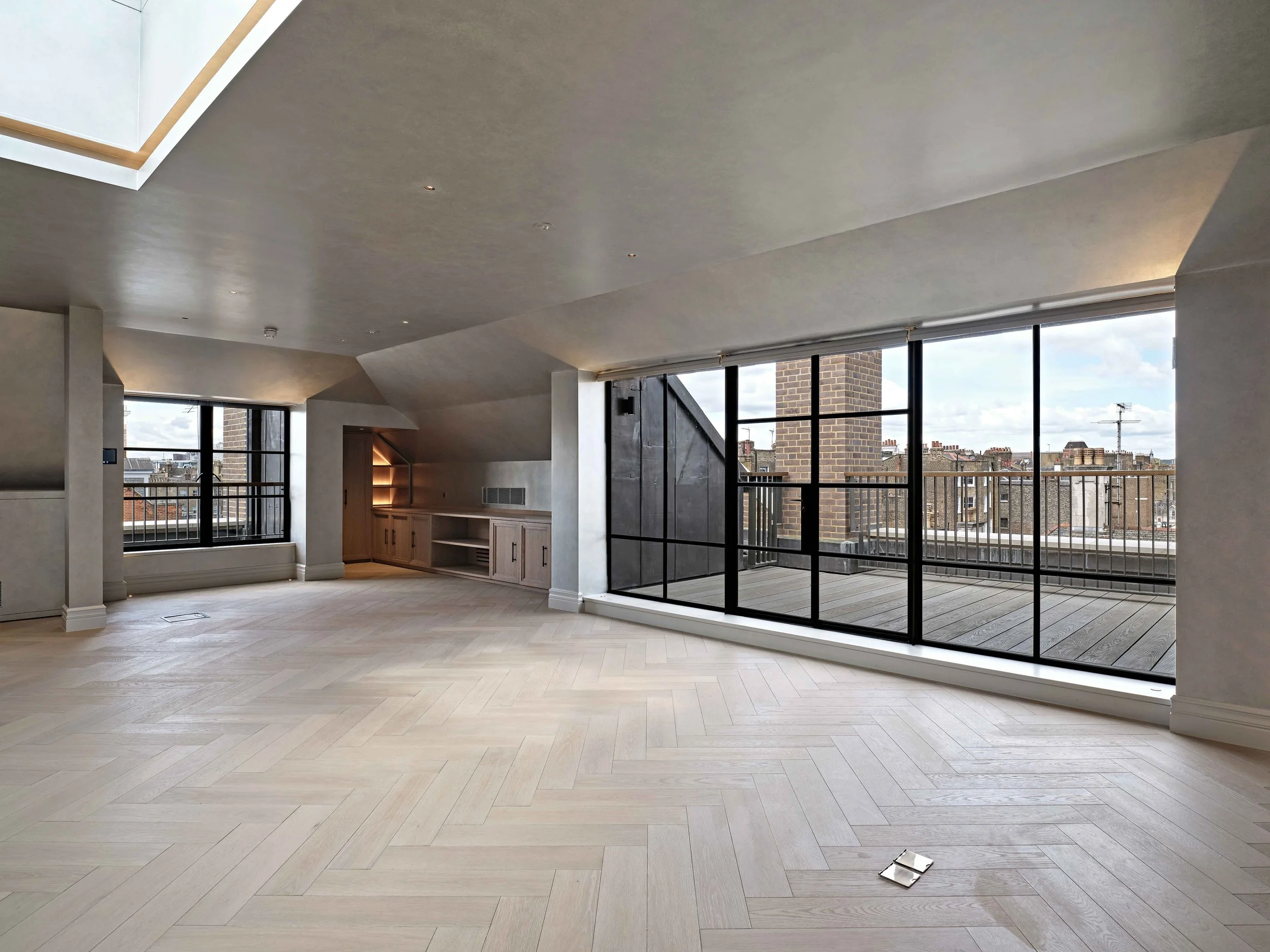 An empty modern living room with light-colored wooden flooring, large floor-to-ceiling windows showing a city view, and a balcony with a black railing. The ceiling has recessed lighting and a skylight.