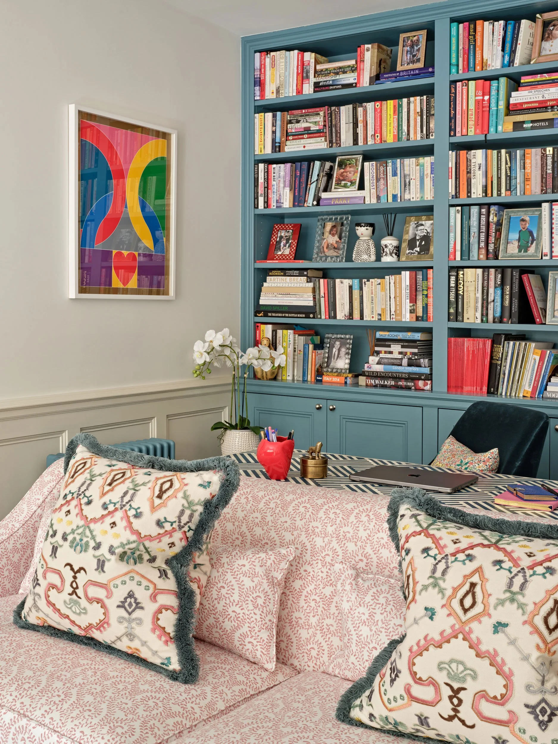 Living room with a pink patterned couch, decorative pillows, and a dark green chair. A table in front has a laptop, notebooks, and a small decorative item. Blue bookshelves filled with books and framed pictures are against the wall, with floral arran