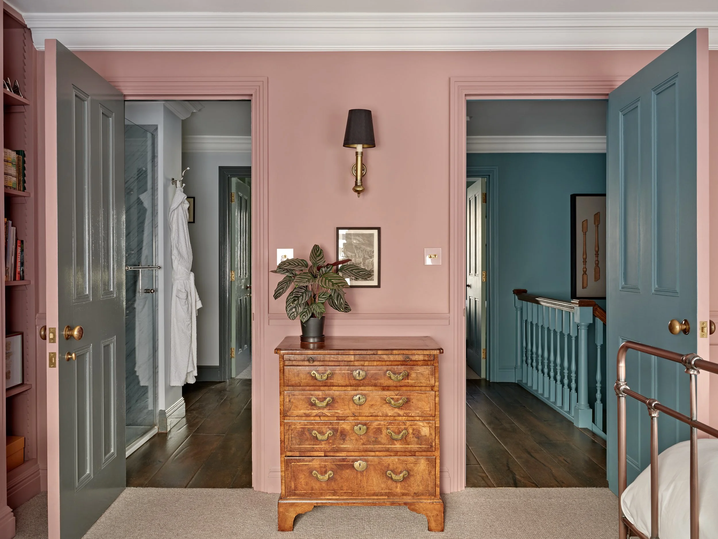 View of a room with pink walls, a wooden dresser in the center, a plant on top of the dresser, and two doorways with blue and gray doors leading to other rooms. There is a black wall sconce above the dresser.