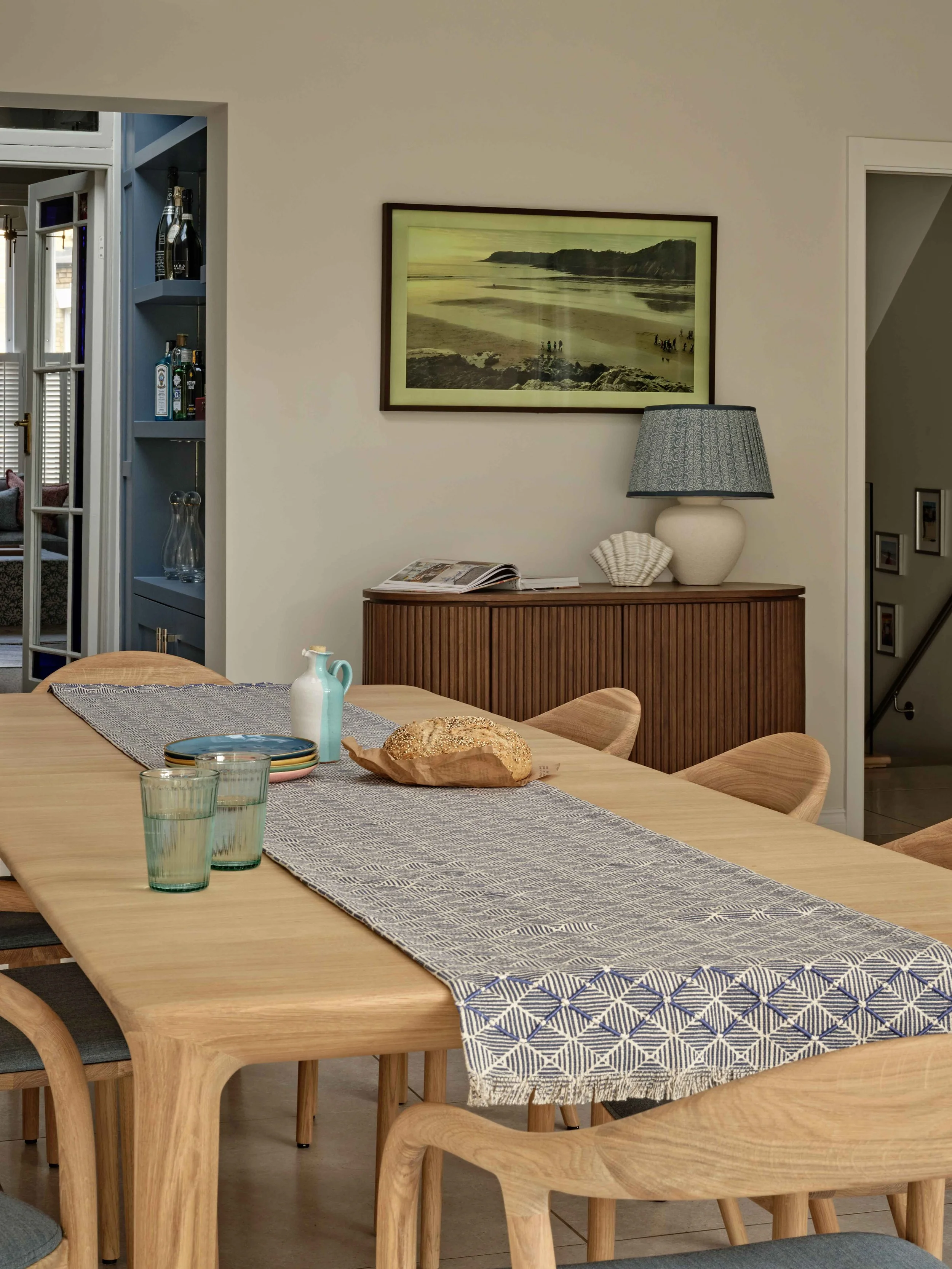 A dining room with a wooden table covered by a patterned table runner, set with glasses, a loaf of bread, and small pitchers. There are wooden chairs around the table, and a sideboard with a lamp, shells, and magazines. A framed beach photograph hang