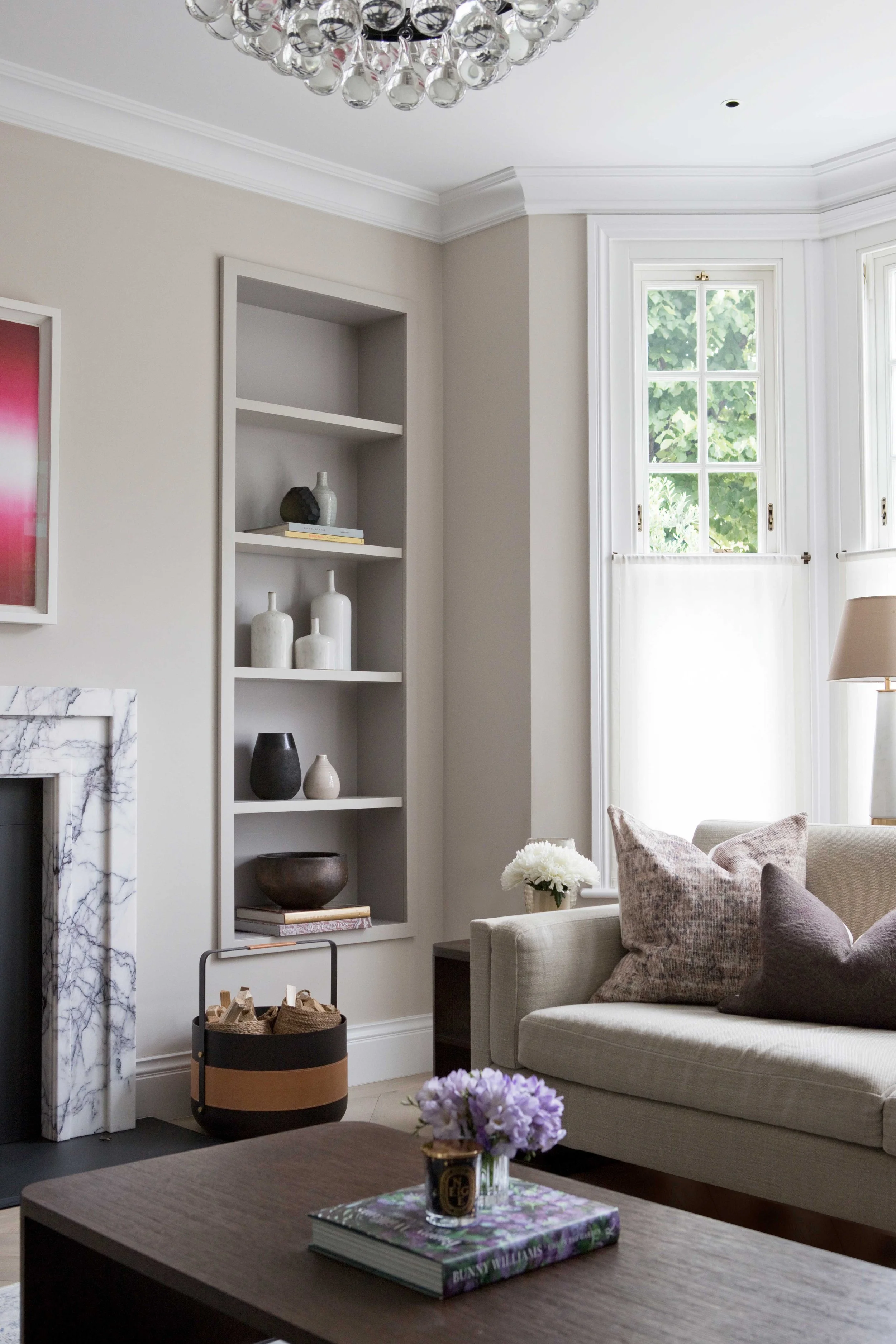 A cozy living room with a beige sofa, decorative pillows, a coffee table with a book, a small vase with purple flowers, a white bookcase with vases, and a fireplace with a marble surround. Large windows let in natural light, and a chandelier hangs fr